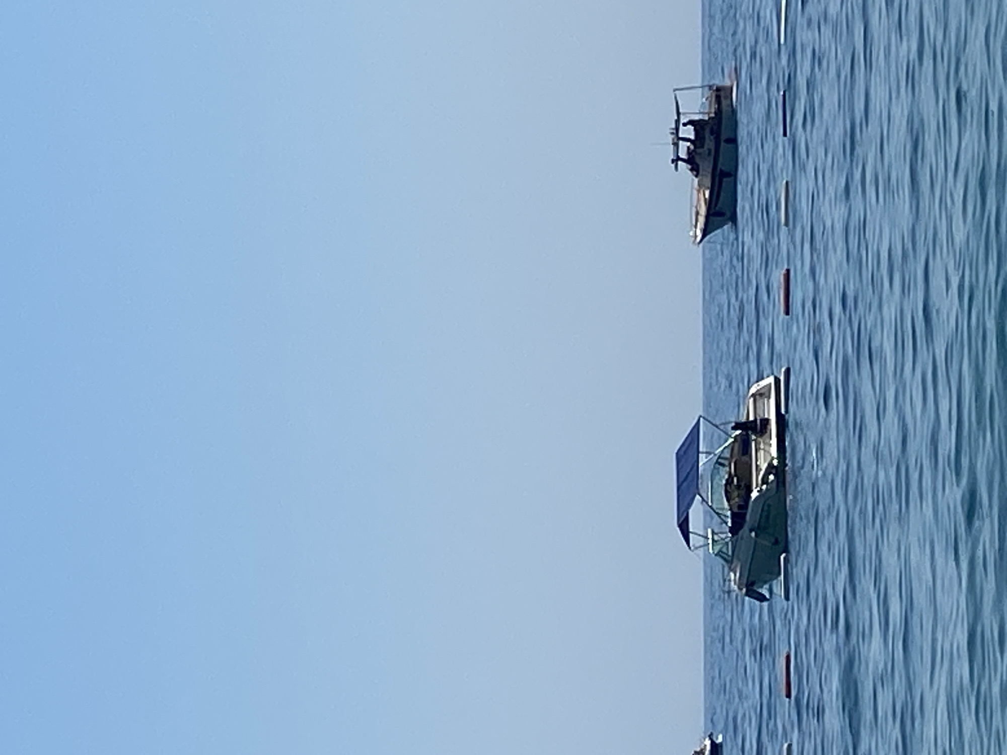 The image shows a calm, blue sea under a clear sky. Several boats are visible on the water. In the foreground, a light-colored speedboat with a blue canopy is anchored. Further out, another, larger boat, also with a canopy, is moving. A series of buoys mark a section of the water. The scene is set against the backdrop of a bright, cloudless sky. The location is Prijevor, Montenegro.