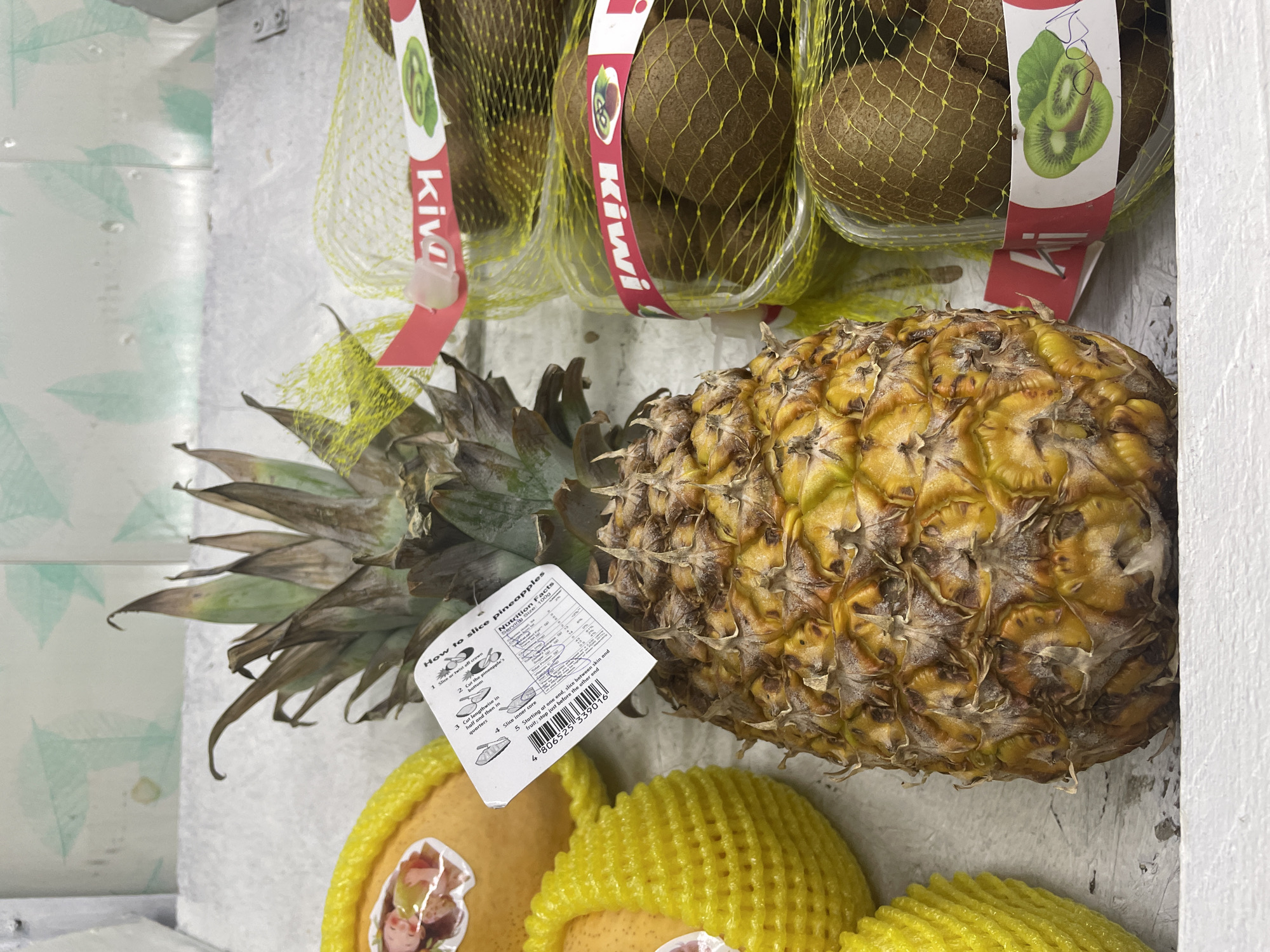 A close-up, eye-level shot captures a pineapple and several packaged kiwis displayed in what appears to be a market or grocery store. The pineapple, prominently featured in the foreground, is a vibrant golden yellow with a textured, spiky rind and a green, leafy crown. A white paper label with instructions on how to slice pineapples and nutritional information is attached to the pineapple. To the right of the pineapple, multiple kiwis are visible, each contained within a yellow mesh bag and secured with a red and white branded label that reads "Kiwi" and features an illustration of sliced kiwis. To the left of the pineapple, several yellow fruits, possibly pears or apples, are also wrapped in yellow mesh netting and have small stickers on them. The background consists of a white shelf or display surface with a light-colored, textured wall behind it, featuring a subtle green pattern. The lighting is even, suggesting indoor illumination. The image does not contain any discernible elements that would indicate its specific location within the city of Almaty, Kazakhstan.