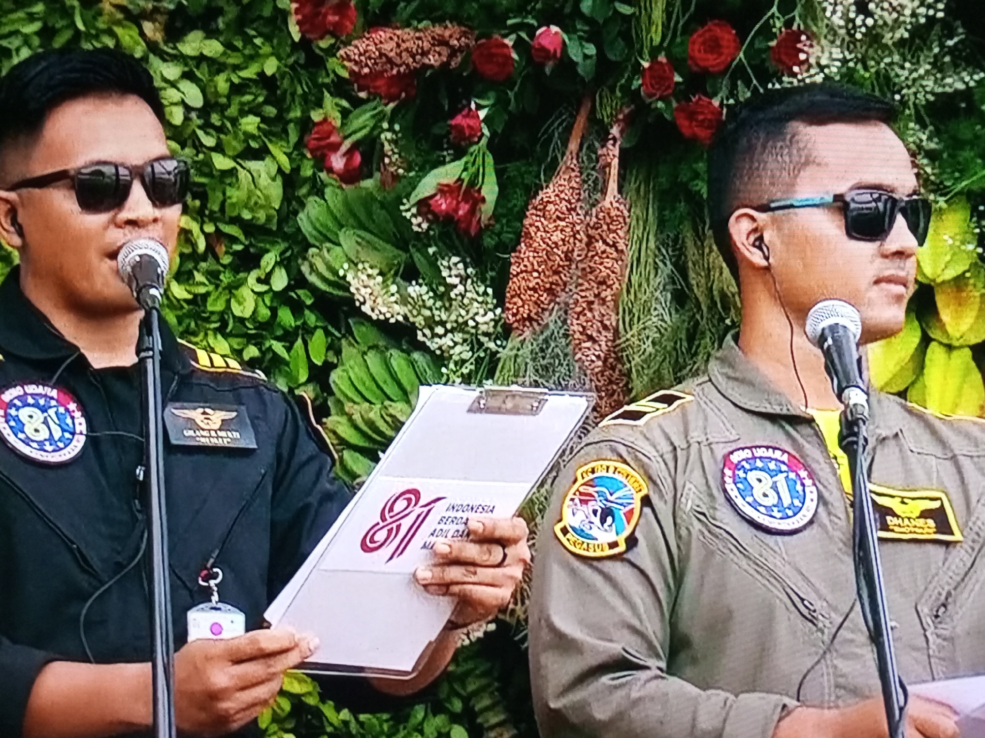 Two men in military-style uniforms stand in front of a backdrop of lush green foliage and red and pink flowers. The man on the left, wearing a black uniform with a circular patch on his left breast featuring the number "81" in blue and red, speaks into a microphone. He wears dark sunglasses and a lanyard with a white tag around his neck. He holds a clipboard with a white surface and a red graphic on it, which appears to be the number "81" intertwined with text that includes "INDONESIA". The man on the right, wearing a khaki-colored uniform with similar patches, also stands near a microphone. He is also wearing sunglasses and an earpiece. The background appears to be an outdoor setting. While the image does not explicitly show the city of Banyudono, Indonesia, the presence of Indonesian language on the clipboard suggests a connection to Indonesia.