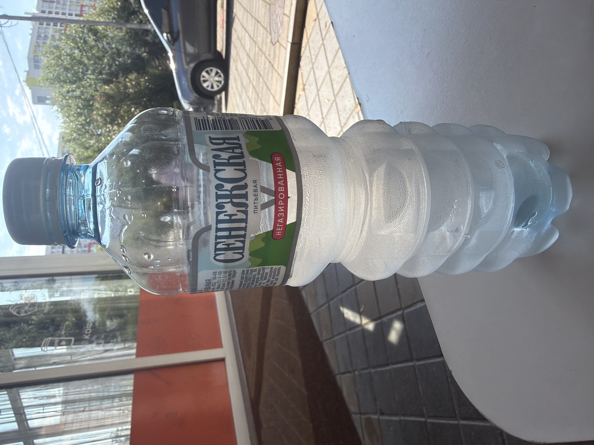 The image shows a plastic bottle of "СЕНЕЖСКАЯ ПИТЬЕВАЯ" (Senezhskaya Pitievaya) water, which is described as "НЕГАЗИРОВАННАЯ" (still) on the label. The bottle is mostly full of water and has condensation on the outside, suggesting it is cold. It is placed on a white surface, possibly a table, outdoors. In the background, there is a tiled walkway, a building with a bright orange facade, and a parked car. The scene appears to be set during the day, with sunlight casting shadows. The overall setting suggests a casual outdoor environment, perhaps a cafe or restaurant patio in Sochi, Russia. The focus is on the water bottle, with the background providing context to its placement. There is no visible activity or event taking place, other than the bottle resting on the surface. The text on the label is in Russian and indicates the brand, type of water, and some production details.