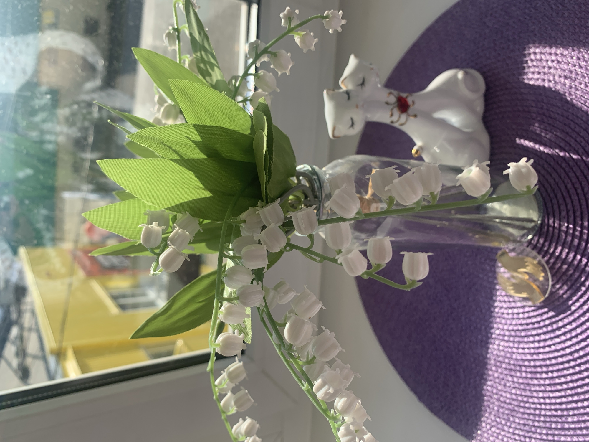 A glass vase containing a bouquet of lily of the valley is resting on a windowsill in what appears to be a private residence. A white porcelain figurine of two cats is positioned behind the vase on a circular purple placemat. Outside the window, a yellow object can be seen. The location is Chaikovsky, Russia.