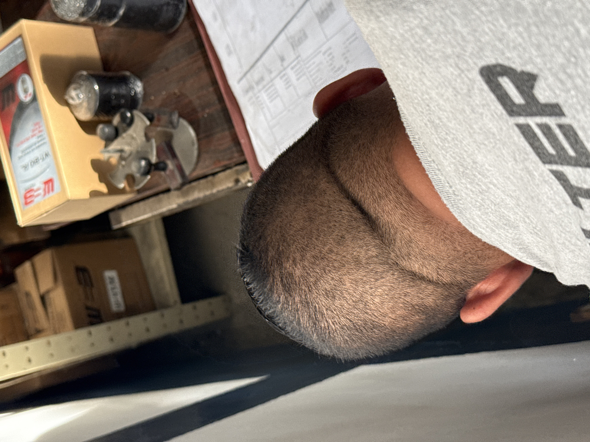 The image shows a close-up of a person's head with very short, dark hair. Part of an ear is visible. The person is wearing a gray shirt with a partial view of a large, dark logo or lettering. Next to the person's head, there's a collection of objects on what appears to be a wooden surface. These include a box with red lettering, black cylinders, and a small, metal multi-pronged tool. Below this, a shelf contains several cardboard boxes. The location is unknown, and no information relates this to Tinaquillo, Venezuela.