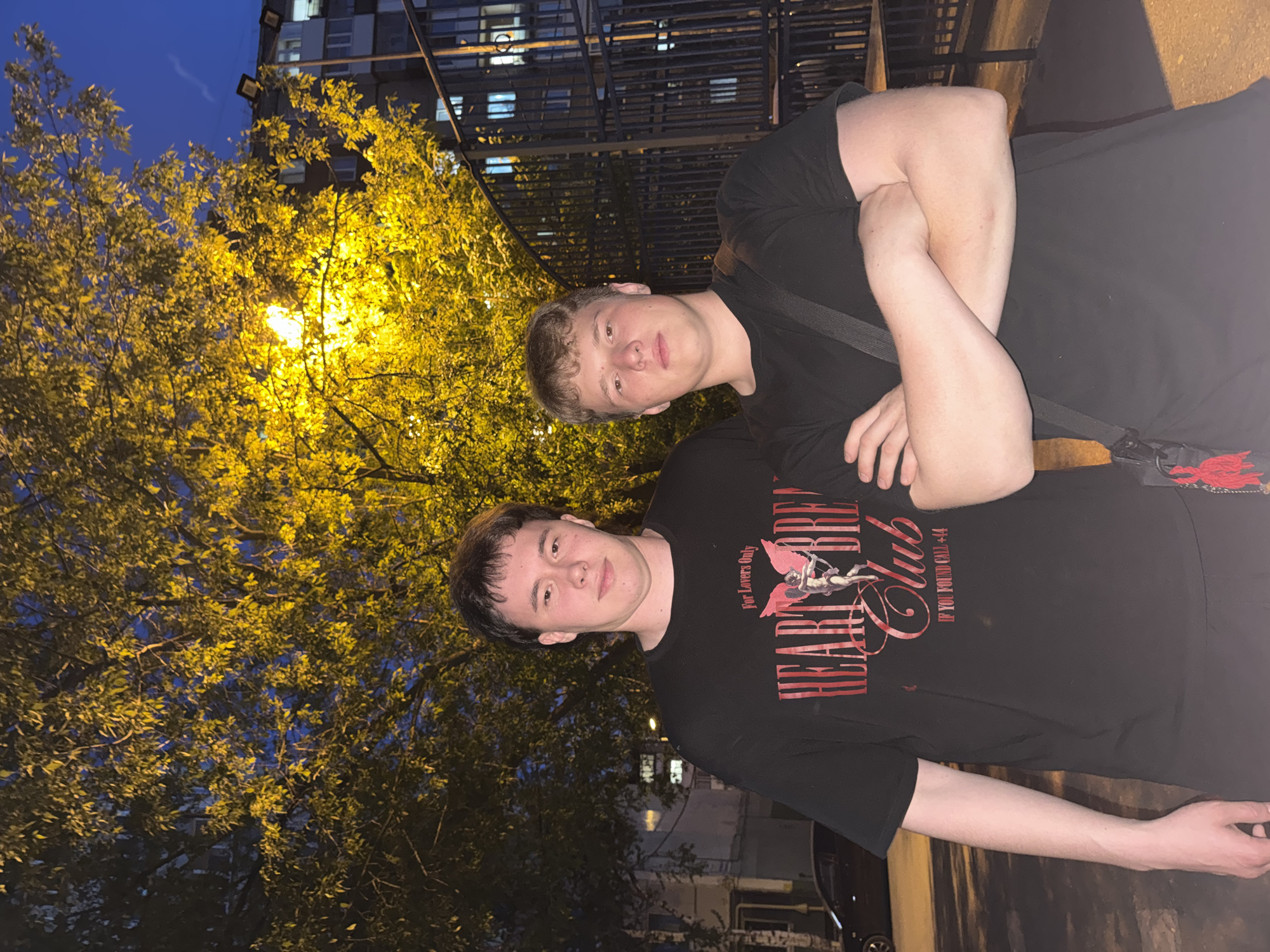 Two Caucasian males are posing for a photograph at night in Perm, Russia. The foreground shows two males. The male in the foreground is wearing a black t-shirt with an image on it and his arm extended toward the right of the frame. Behind him, another male is posed with his arms crossed, wearing a black t-shirt. Behind the males, there is a large tree with yellow leaves that is illuminated by a street lamp. In the background, there are buildings and a metal structure. The scene is dimly lit.