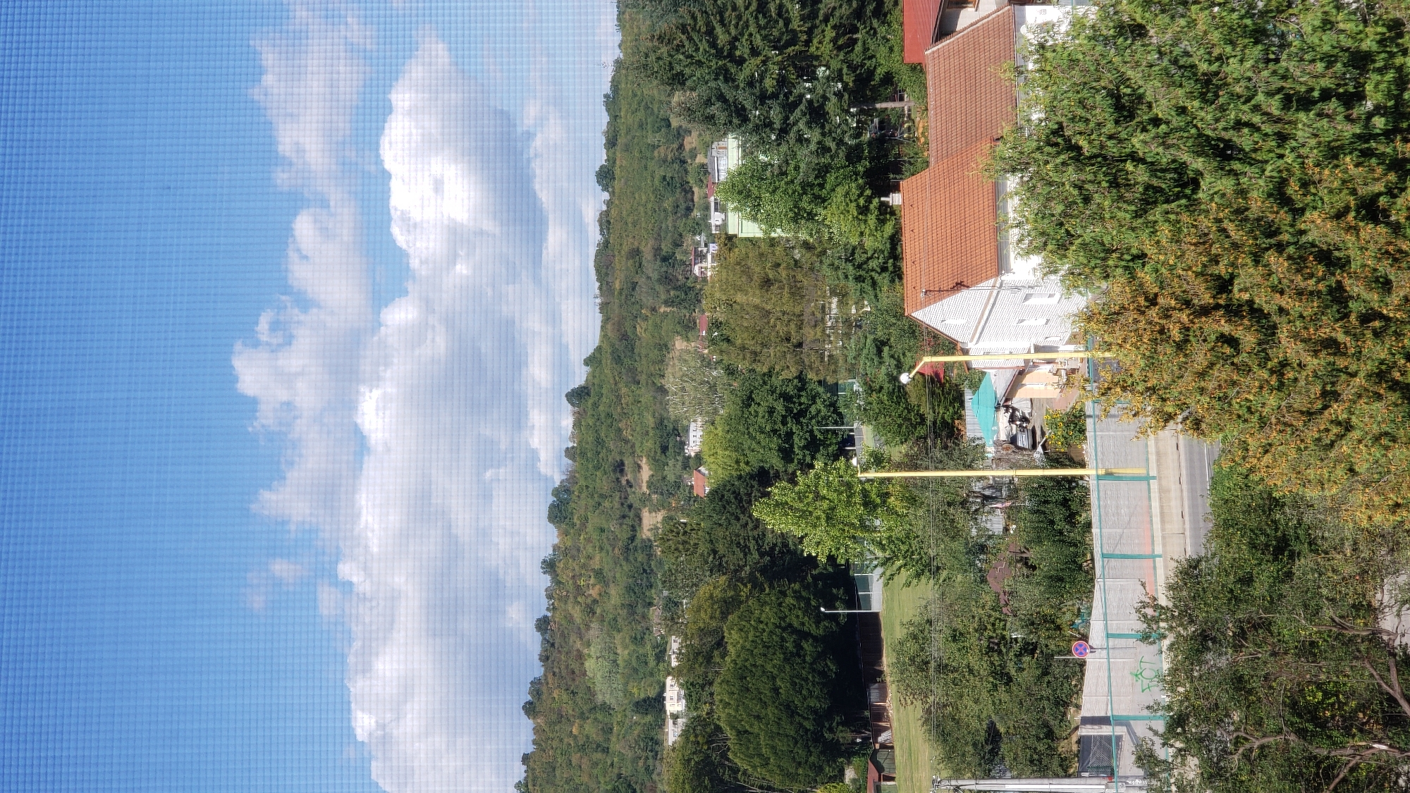 The image depicts a view of a residential area with a hilly, tree-covered landscape in the background. In the foreground, there are houses with red tiled roofs, lush green trees, and a concrete barrier along a road. A yellow pole with a light fixture stands prominently in the mid-ground. The sky is blue with scattered white clouds. Based on architectural style and vegetation, the location is consistent with a suburban or semi-rural area. The scene appears to be captured during daylight hours on a clear day.