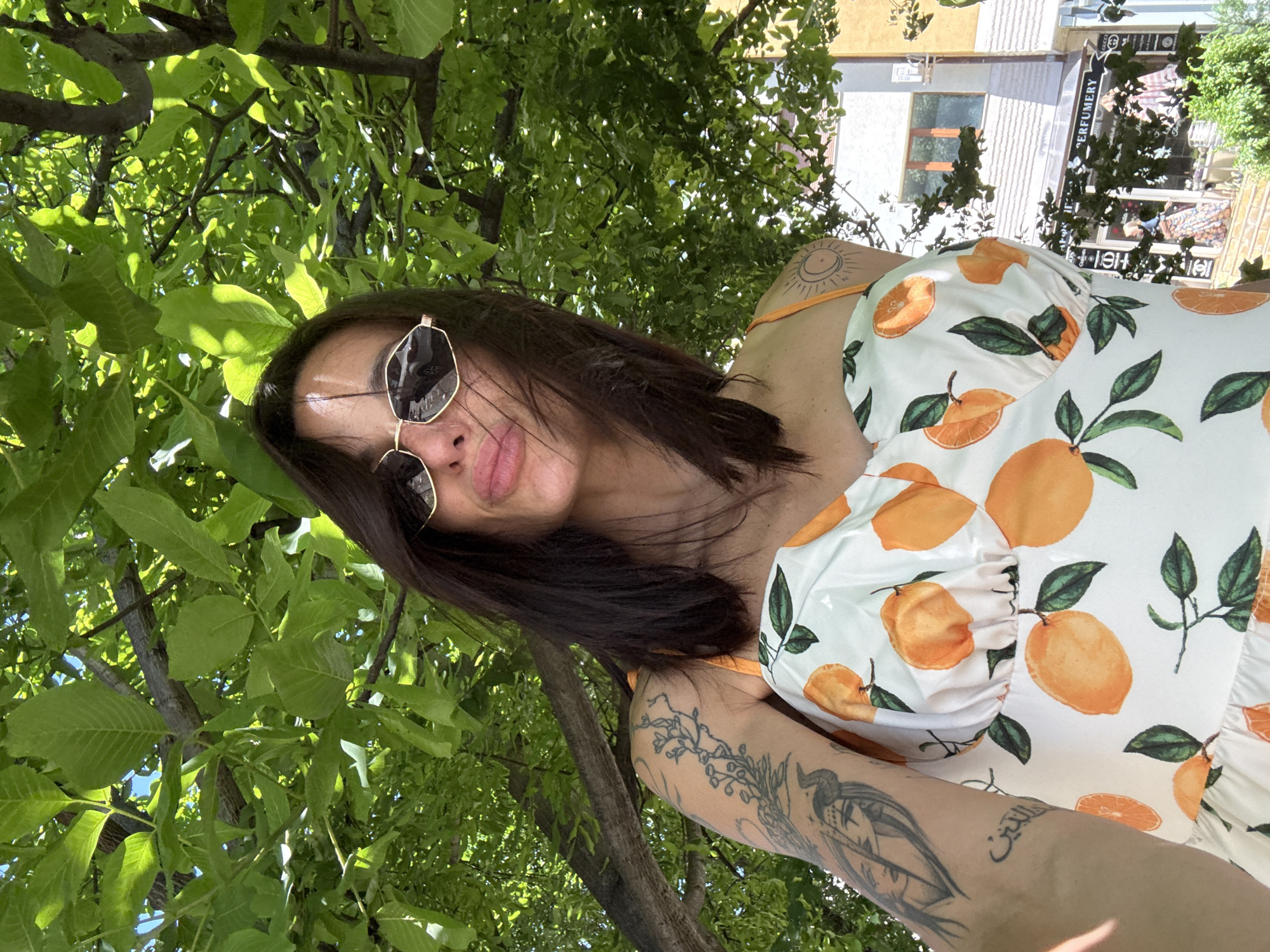 A woman with dark hair and sunglasses is taking a selfie outdoors, with green leaves from a tree in the foreground and a street scene in the background. She is wearing a white dress with an orange fruit print. Tattoos are visible on her left arm and shoulder. In the background, buildings line a street, with signage including "PERFUMERY" and "GUCCI". The woman appears to be on a sunny day, as indicated by the lighting.