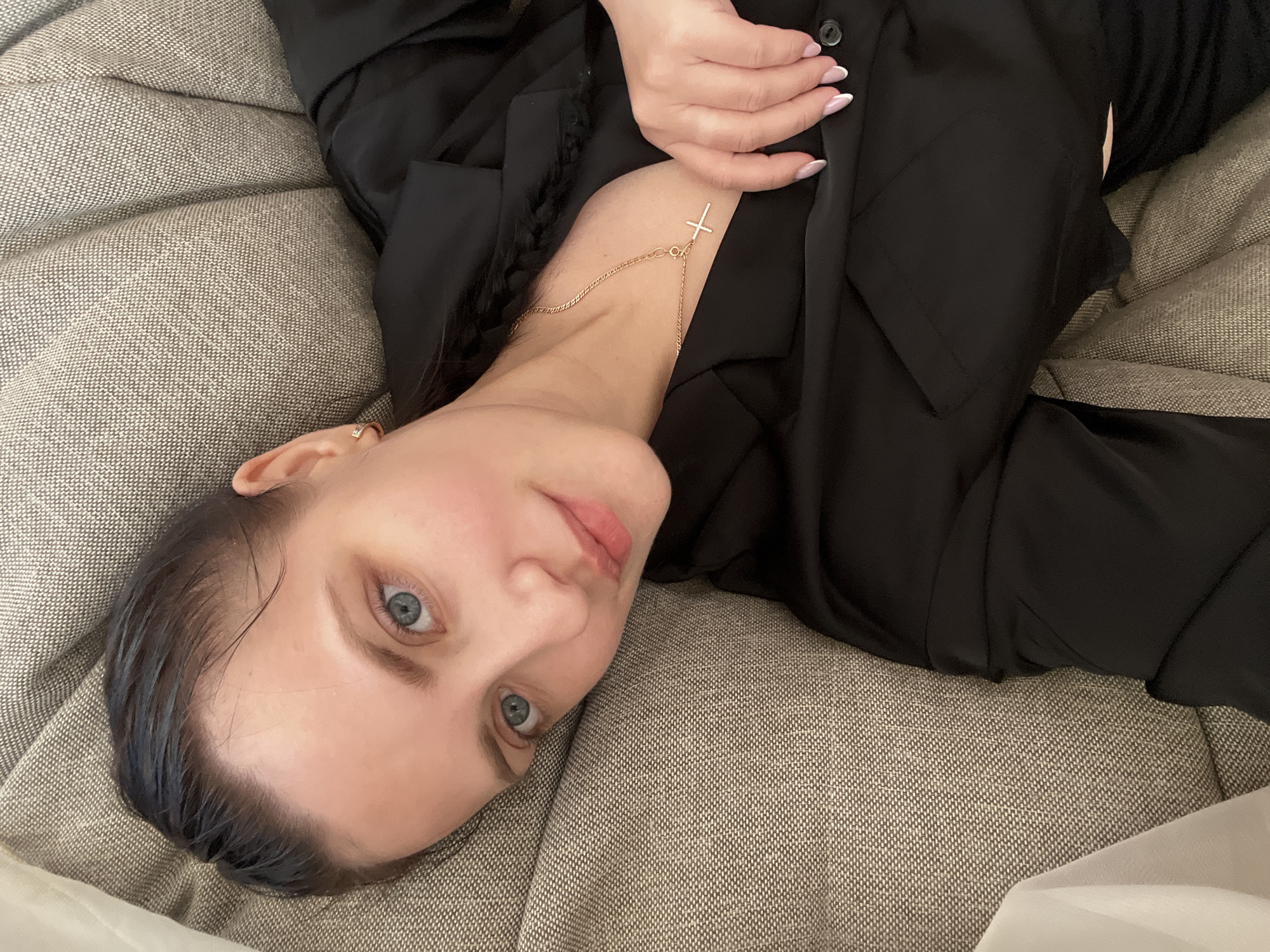 An overhead shot shows a woman lying on her back on a textured, light brown-grey upholstered surface, likely a sofa or bed. Her head is angled to the left, and she looks directly at the camera with clear blue eyes. Her dark hair appears slightly wet or slicked back, revealing her forehead and hairline. She has a fair complexion and light pink lip color.
She is wearing a plain black long-sleeved garment, possibly a shirt or jacket, which covers most of her upper body. Around her neck is a delicate gold-colored chain necklace with a small, inverted cross pendant hanging from it. One of her hands, adorned with a French manicure, rests near her shoulder on the black fabric. A small ear cuff or earring is visible on her left ear.
The furniture has a woven fabric texture, providing a neutral background. The woman's pose suggests a relaxed state, possibly a casual moment or a planned portrait. There is no visible text in the image. The setting appears to be indoors.