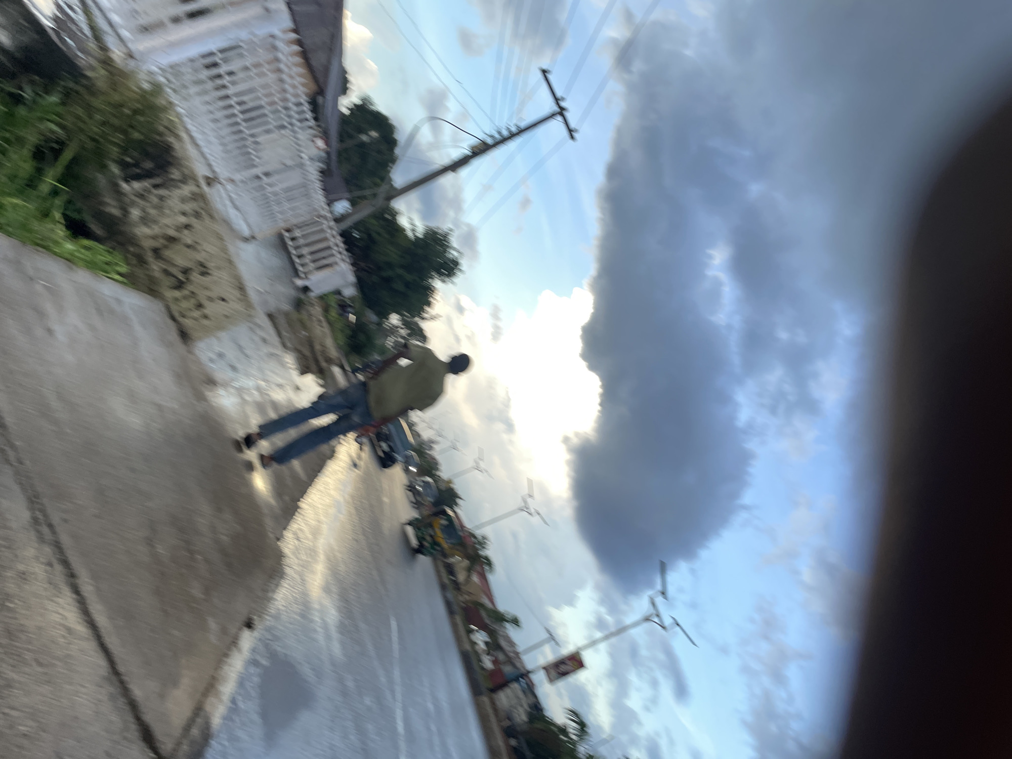 The image shows a street scene, potentially in Jos, Nigeria. A person in jeans and a light-colored shirt is walking on a wet sidewalk, reflected in the pavement. Beyond the sidewalk, there are buildings with white fences and lush green vegetation. A utility pole with power lines stands prominently in the frame, with a cloudy blue sky above. The image is somewhat blurry and appears to be taken at an angle, possibly upside down. There is a sense of urban environment mixed with natural elements.