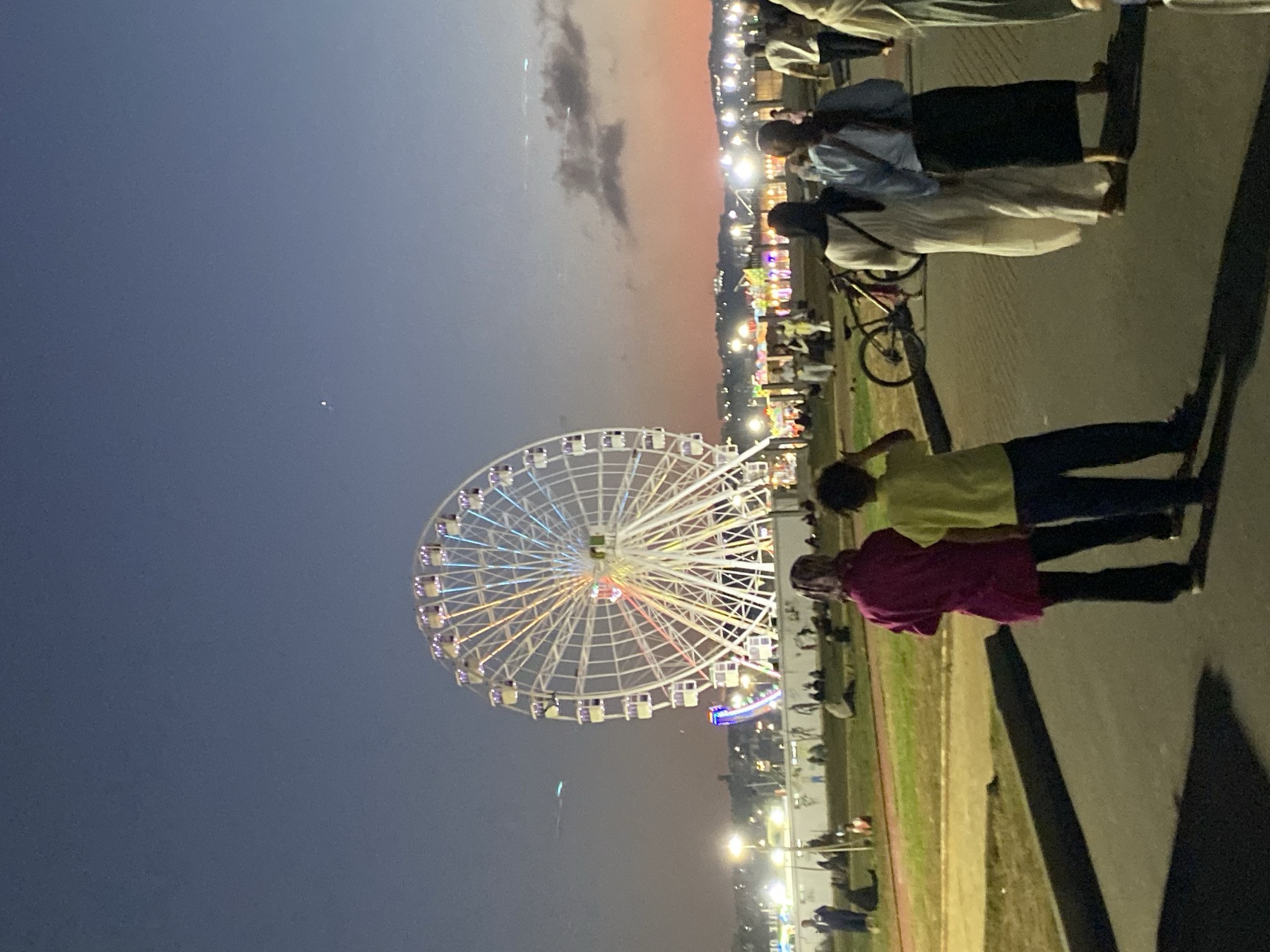 In Hussein Dey, Algeria, a large Ferris wheel illuminated with colorful lights dominates the twilight scene. A paved walkway leads into the frame, where several people are walking. In the foreground, a person in a magenta top and black pants stands with their back to the camera, next to another person in a yellow shirt and blue pants. To the right, three figures, two women wearing hijabs and long dresses, and one woman in a light-colored headscarf, walk away from the viewer. A bicycle is parked nearby. In the middle ground, a grassy area is populated with more people sitting and standing, and further in the background, a collection of amusement rides and stalls are brightly lit. The sky transitions from a deep blue at the top to a warm orange and pink horizon.