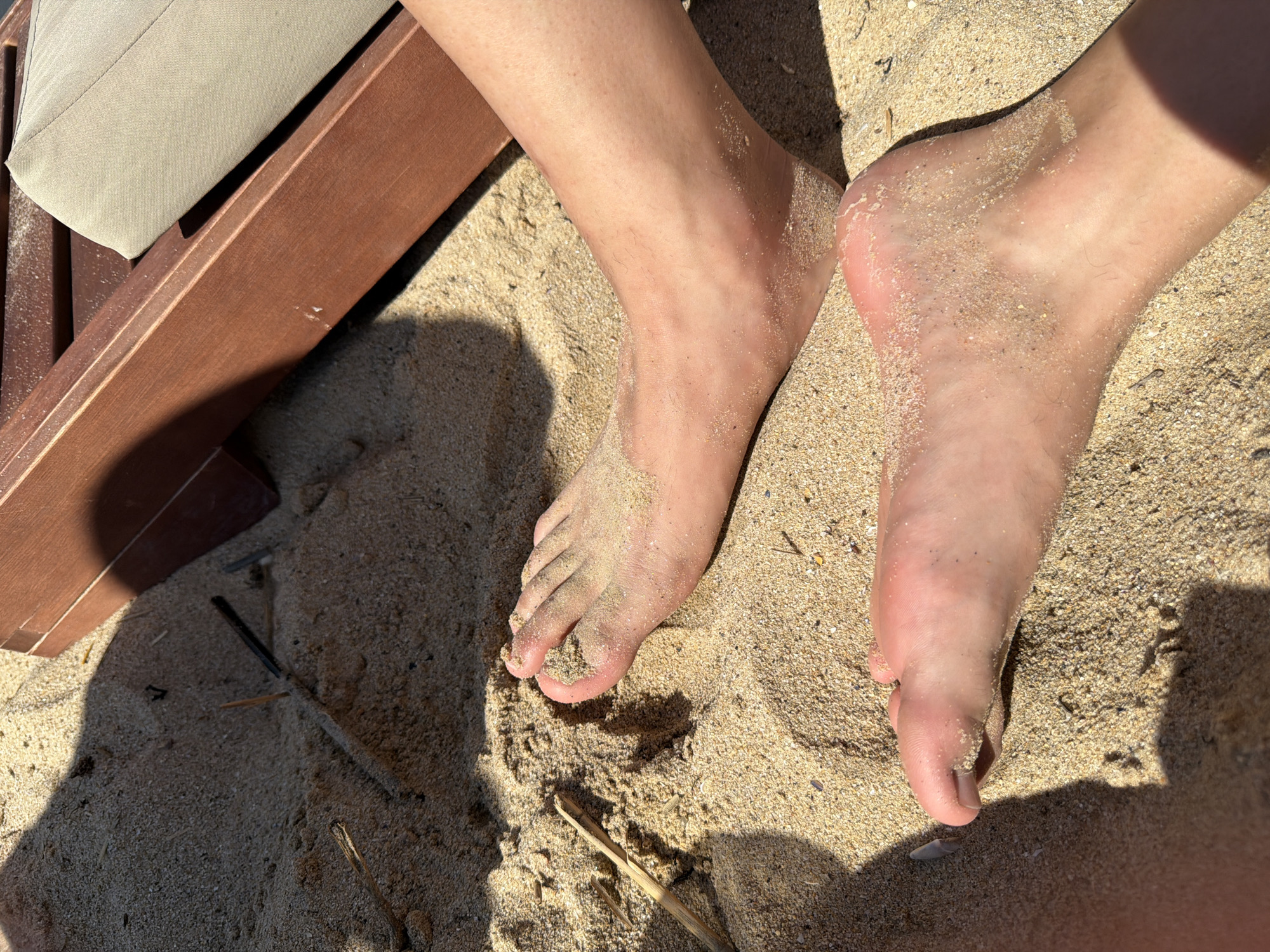 The image depicts a close-up view of two human feet partially covered in sand, resting on a sandy surface. A portion of a wooden beach lounger with a beige cushion is visible in the upper right quadrant of the image, suggesting an outdoor, possibly beach, setting. The lighting indicates a sunny day, casting shadows on the sand. The sand itself appears coarse with small pebbles and fragments of dried plant material scattered throughout. The feet appear to be clean with no visible injuries or adornments. The overall scene is static and does not convey any specific actions or interactions. The location is identified as Varna, Bulgaria.