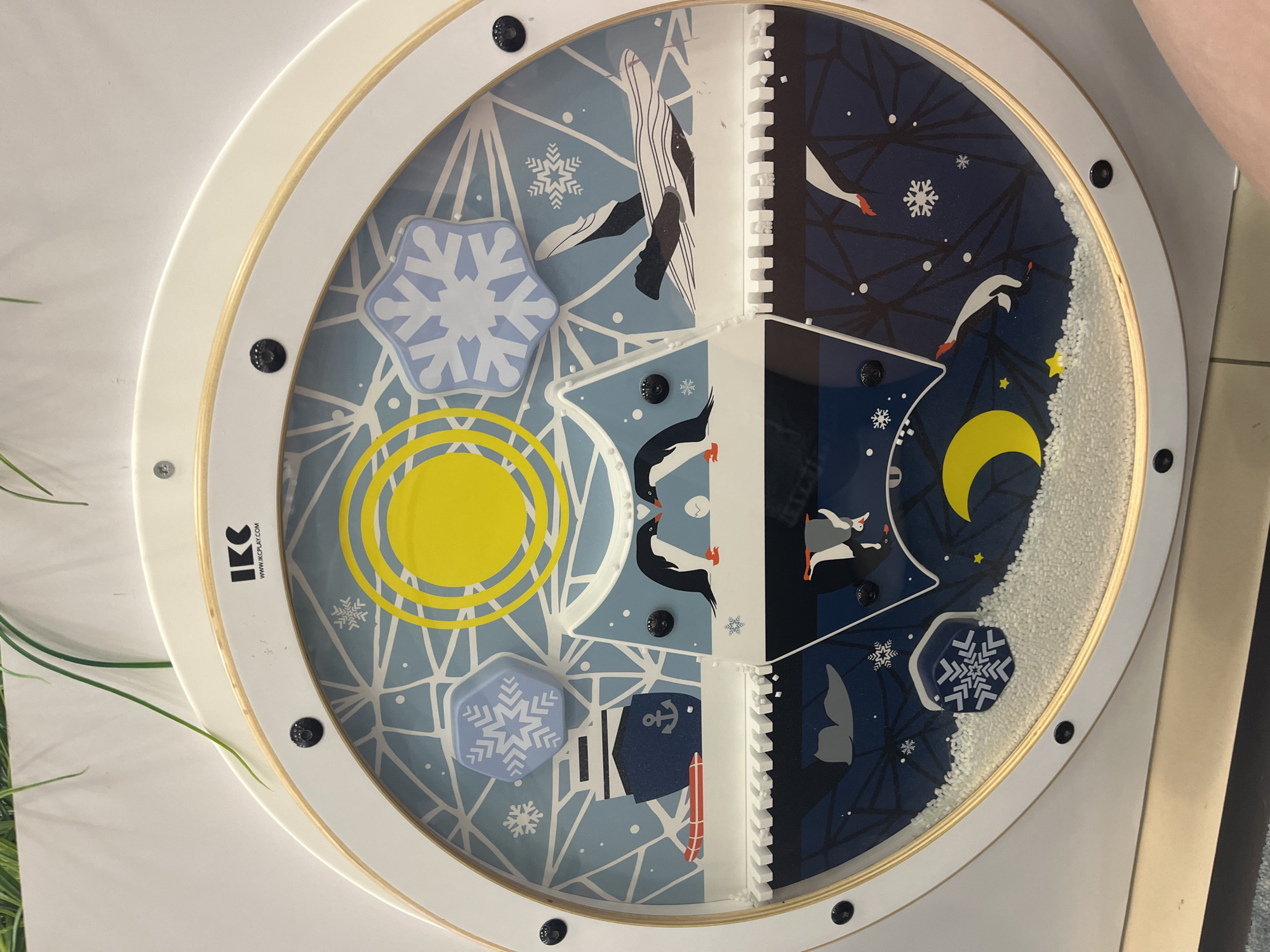 The image displays a circular, wall-mounted interactive play panel, approximately 80-90 cm in diameter, located in the city of Unknown, Unknown. The panel has a white outer frame with an inner visible wooden ring and is secured by multiple black fasteners around its circumference. The top section of the frame features the black logo "IKC" and the website "WWW.IKCPLAY.COM".
The panel's interior is divided into two primary themed sections by a horizontal, irregular line suggesting an ice edge or landmass.
The upper section depicts a daytime, above-water Antarctic or Arctic scene. The background is light blue with white geometric patterns resembling ice floes and scattered white snowflakes. A large yellow sun is prominent at the top center. A light blue hexagonal piece with a white snowflake design is positioned in the upper left quadrant. To the left, a dark blue ship with a white anchor is shown alongside a red sled. In the center, two penguins stand facing each other, their beaks almost touching, with a small white egg between them. To the right, a large black and white whale is depicted partially submerged.
The lower section illustrates a nighttime, underwater or subterranean scene. The background is dark blue with white geometric patterns and scattered white snowflakes. A yellow crescent moon and several small yellow stars are visible in the lower left. Two penguins are shown, one diving headfirst into the water and another inverted. A grey whale tail is visible near the center. The very bottom portion of this section is filled with a layer of white granular material, resembling snow or small beads. A dark blue hexagonal piece with a white snowflake design is partially embedded in this material in the lower left.
The panel is mounted against a plain white wall, with a few thin green plant fronds visible extending into the frame from the top edge. The bottom right corner of the image shows a partial view of a light-colored floor and a person's bare foot.