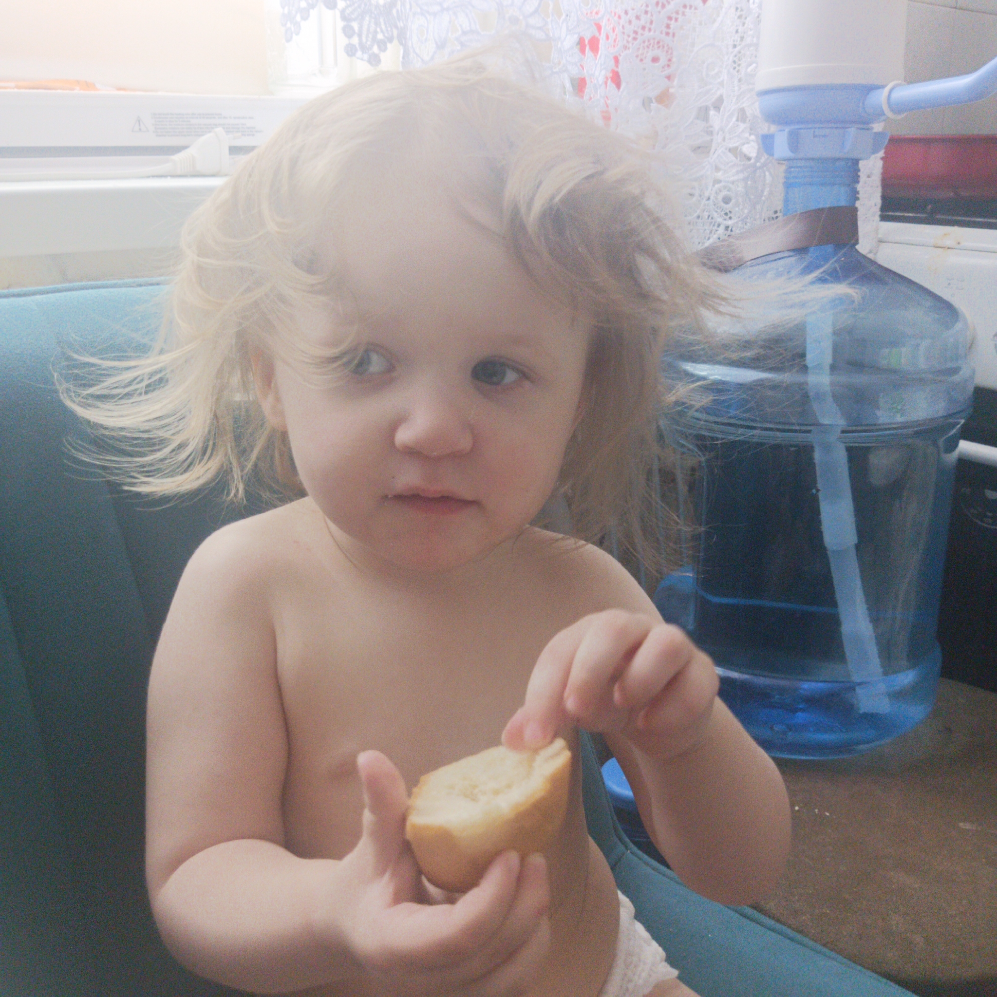 A young child with blonde, windblown hair sits in a blue upholstered chair. The child is holding a piece of bread in their hands. A large blue water bottle with a white pump mechanism sits on a surface to the right of the child. A window with a white lace curtain is behind the water bottle. The background appears to be a kitchen setting. The image was taken in Chaikovsky, Russia.