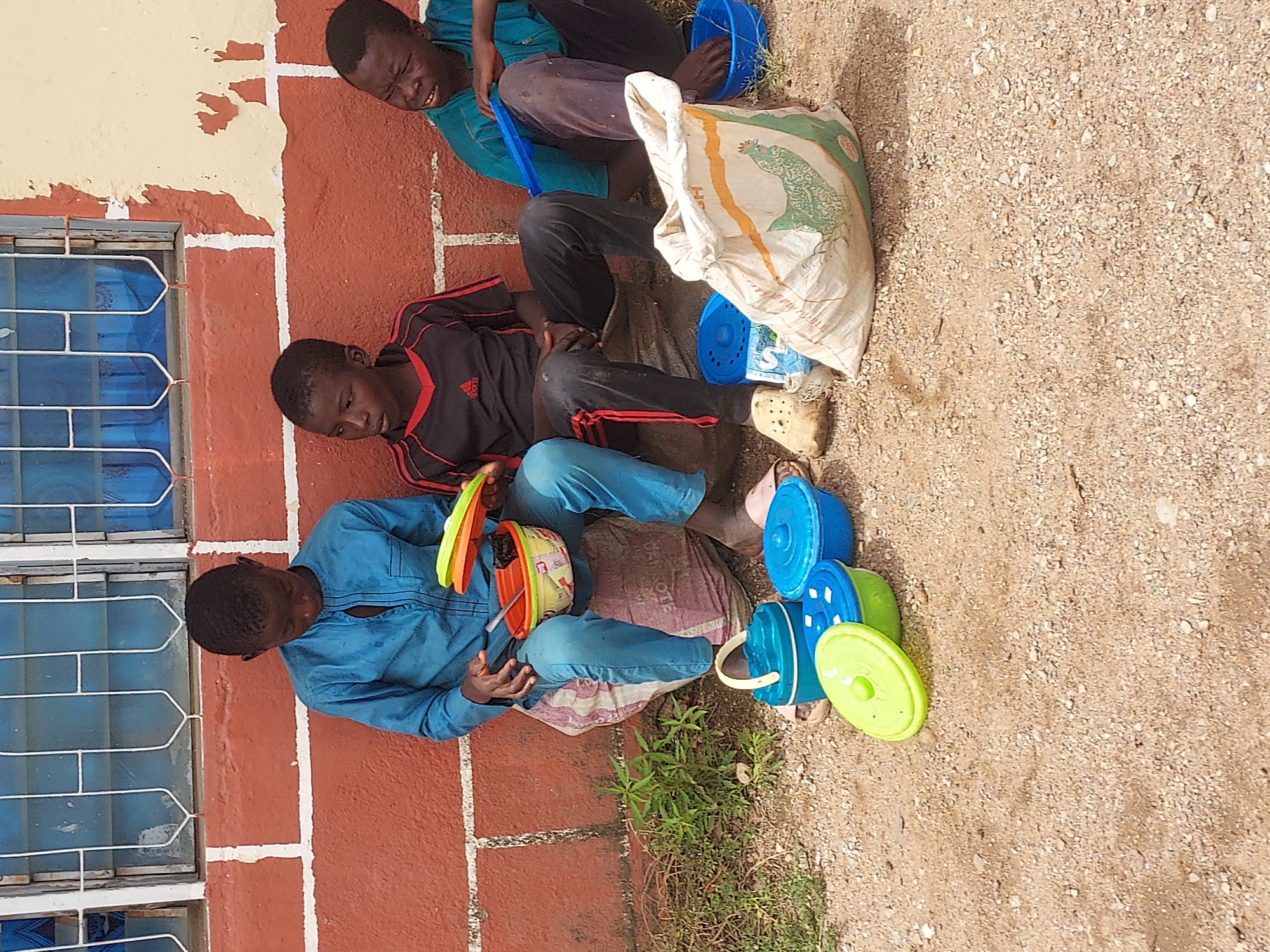 Three male individuals are seated against a red brick wall in an outdoor setting. The location is inferred to be in Jos, Nigeria. The wall has a window with blue panes and white bars. The individuals are of African descent.
The person closest to the window wears a blue shirt and jeans. They are holding a stack of colorful plastic containers. The second individual wears a dark shirt with red stripes, and dark pants. The third individual wears a teal shirt and dark pants and is holding a blue container.
To the right of the individuals is a large white sack with an image on it, along with several plastic containers of various colors and sizes. The ground appears to be a mixture of dirt and small pebbles. Some small weeds grow near the individuals.