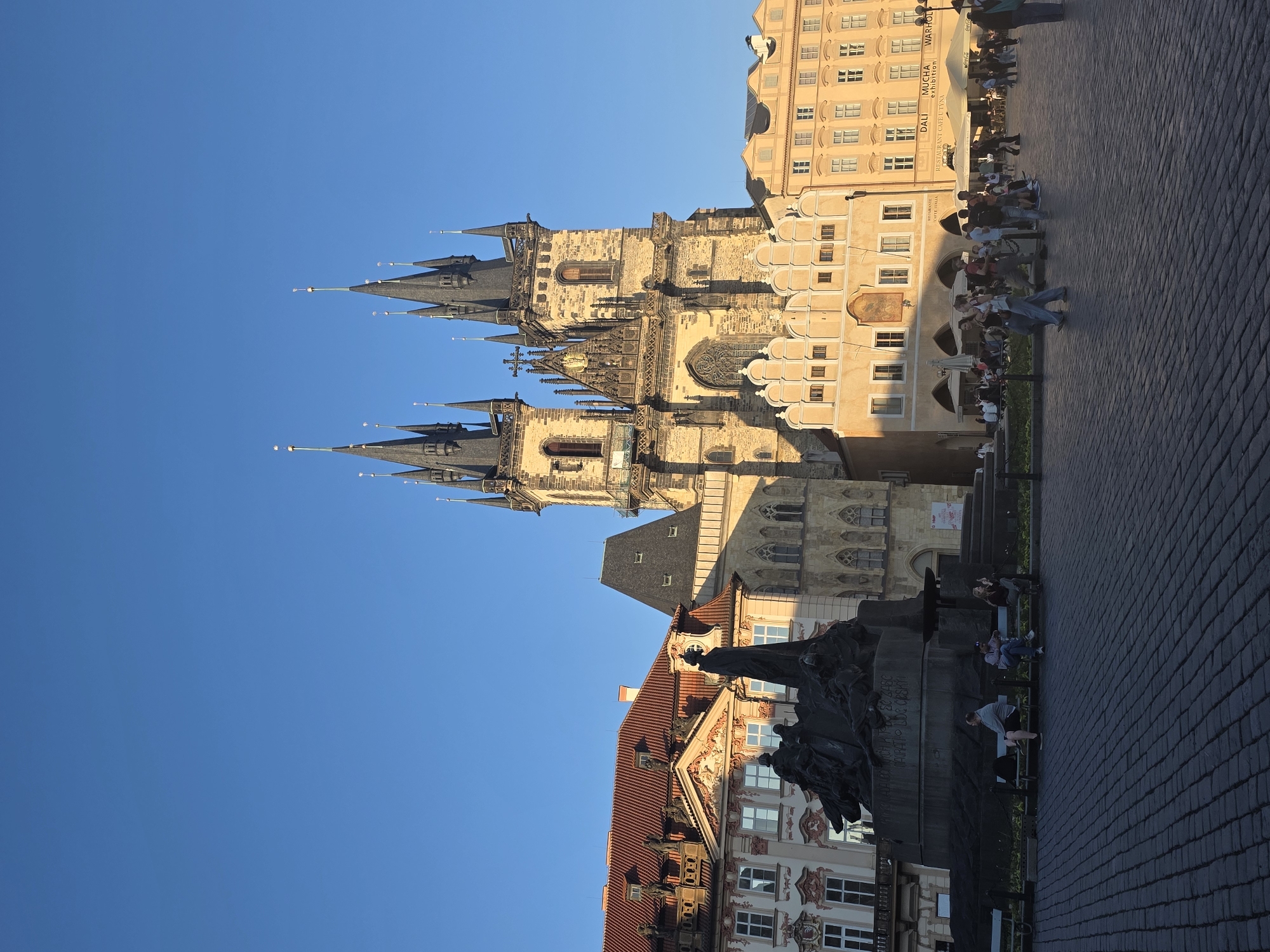This image captures a bustling late afternoon or early evening scene in a historic European city square, likely Prague's Old Town Square, under a clear, deep blue sky. The warm, golden light of the sun casts long shadows, indicating the "golden hour."
The most prominent feature is the gothic Church of Our Lady before Týn, which dominates the mid-ground. Its multiple dark, pointed spires rise majestically, and its intricate stone facade is bathed in sunlight, revealing detailed arches and windows. Adjacent to the church are other historical buildings with a warm, lighter-colored facade, housing ground-level establishments.
In the foreground and to the left, a large, dark bronze statue, identifiable as the Jan Hus Memorial, is visible. Several individuals are sitting on benches around its base, some looking at their phones or simply relaxing. Further into the square, other people are seen walking or gathered at outdoor cafes with tables, chairs, and white umbrellas. One building features signs for "DALI MUCHA WARHOL exhibition" and "RESTAURANT CAFÉ U TÝNA." The square itself is paved with cobblestones.
The base of the Jan Hus Memorial displays fragmented text, partially captured by OCR as "CH KITSC ZASC" and "NAIRATH LIDE CESKYI," which are portions of Czech inscriptions. The scene depicts a typical lively urban environment, suggesting a pleasant, relaxed atmosphere.