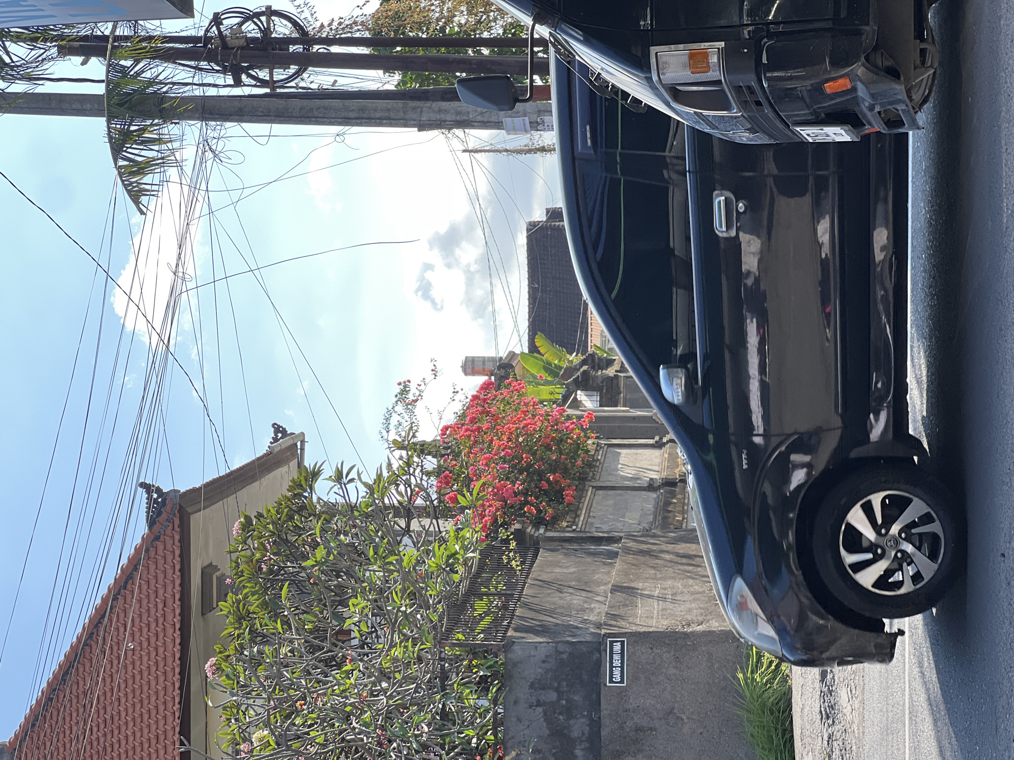 The image shows a street scene in Denpasar, Indonesia, on a sunny day. In the foreground, two black vehicles are parked side by side. The vehicle on the left is a sedan, and the one on the right is a van. Behind the vehicles, a stone wall runs horizontally across the frame, with a metal fence on top. Lush green bushes and vibrant pink bougainvillea flowers cascade over the wall. A sign on the wall reads "GANG DEWI UMA," indicating a street name. In the background, a red-tiled roof of a house is visible, along with a cluster of utility poles and a tangle of overhead electrical wires stretching across the bright blue sky with scattered white clouds. A palm tree branch extends into the frame from the right.