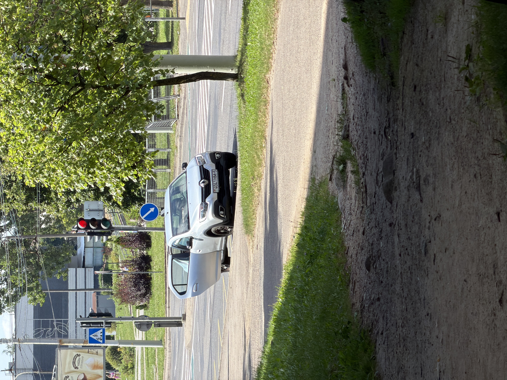 In Minsk, Belarus, a silver Renault Logan is parked on the right side of a road, with its driver's side door open. The car is facing left, towards a pedestrian crossing. Traffic lights are visible, with a red light illuminated. A blue sign indicating a pedestrian crossing is posted on a pole. The scene is captured on a sunny day, with shadows cast across the road and sidewalk. Green grass and trees are present in the background, suggesting a well-maintained urban environment. The road appears to be an intersection with multiple lanes and marked pedestrian and vehicle pathways.