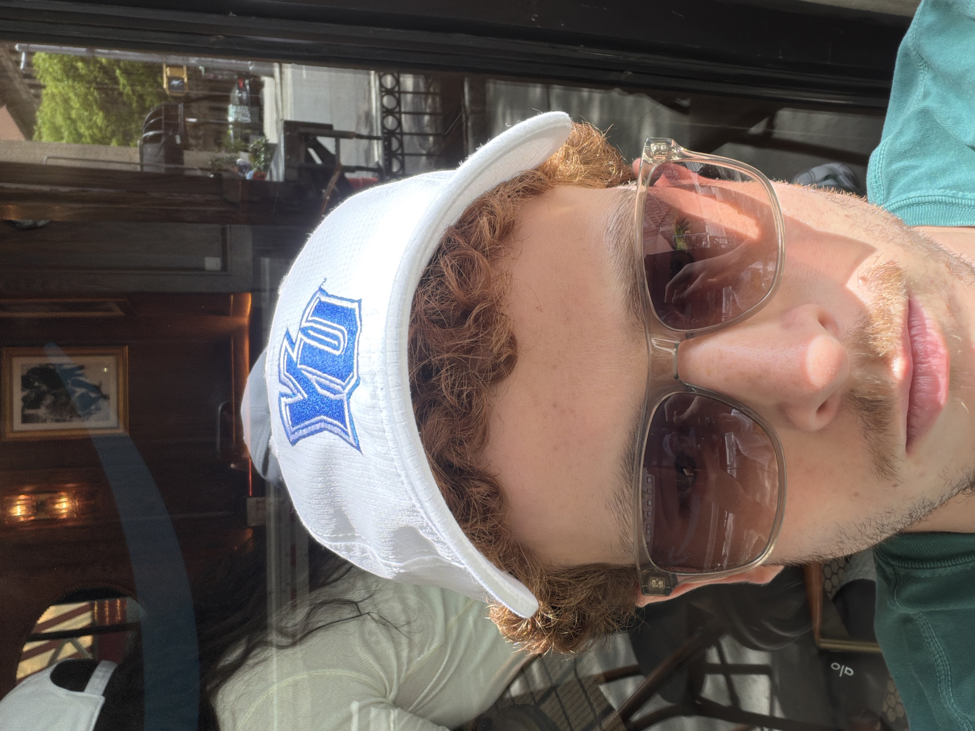 A man is pictured in a close-up selfie. He is wearing a white baseball cap with a blue emblem, tinted sunglasses, and a green shirt. His curly red hair is visible under the hat. Reflections in the glass behind him reveal an interior, possibly a restaurant or bar, with wood paneling and framed artwork. Outside, a city street is visible, with trees and other buildings. Based on the style of the architecture and other cues, the location appears to be in New York City, United States of America.