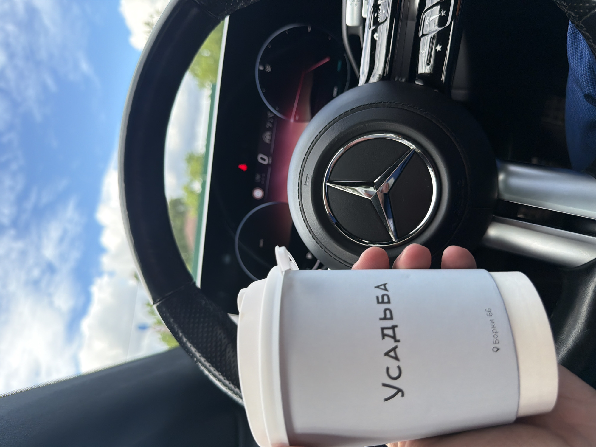 A medium shot captures a hand holding a white coffee cup with the inscription "УСАДЬБА" and "с. Борки 66" (the village of Borki 66), suggesting the drink originates from or is associated with the locality of Borki in Russia. The background features the interior of a vehicle, specifically the steering wheel with the prominent Mercedes-Benz logo, and the dashboard area. The lighting suggests a bright, possibly daytime setting, with the sky visible through the windshield. The image appears to be taken from the driver's seat, with the cup held in front of the steering wheel.