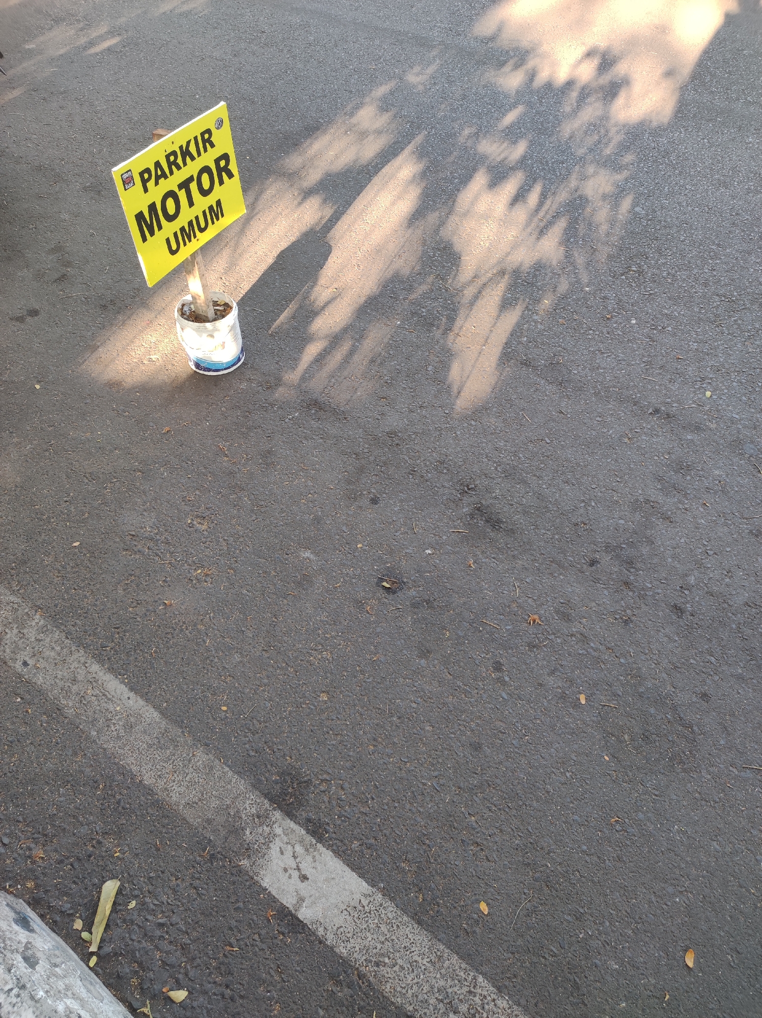 A yellow sign with black lettering reads "PARKIR MOTOR UMUM" which translates to "Public Motorcycle Parking". The sign is attached to a wooden stick and placed inside a white bucket filled with dirt and possibly concrete, serving as a makeshift base. The sign is tilted slightly towards the right. It is located on an asphalt road, with a white painted line indicating a parking or traffic boundary running diagonally across the lower left portion of the image. The scene is illuminated by the sun, casting dappled shadows of tree leaves across the road. The overall environment appears to be an outdoor urban setting, likely a street or parking area. There are no people or vehicles visible in the image. The location is identified as Malang, Indonesia.