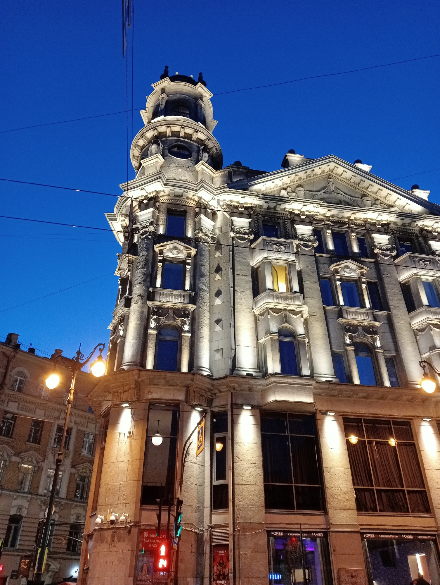 The image shows the corner of a grand, illuminated building in Saint Petersburg, Russia, at dusk. The sky is a deep, clear blue. The building, with its ornate architectural details, is lit by numerous spotlights, highlighting its classical facade. It features columns, decorative friezes, and a prominent turret with a rounded roof topped with small spires. Several windows are visible, some reflecting the blue sky, others showing interior lights. On the street level, there are large windows with dark curtains and signage that reads "FORBS". To the left of the main building, another building with a similar architectural style is partially visible, also illuminated. A street lamp with two glowing bulbs stands in the foreground, casting warm light. Power lines crisscross the sky, a common sight in urban environments. The overall impression is one of an elegant, historic city at the beginning of the evening.