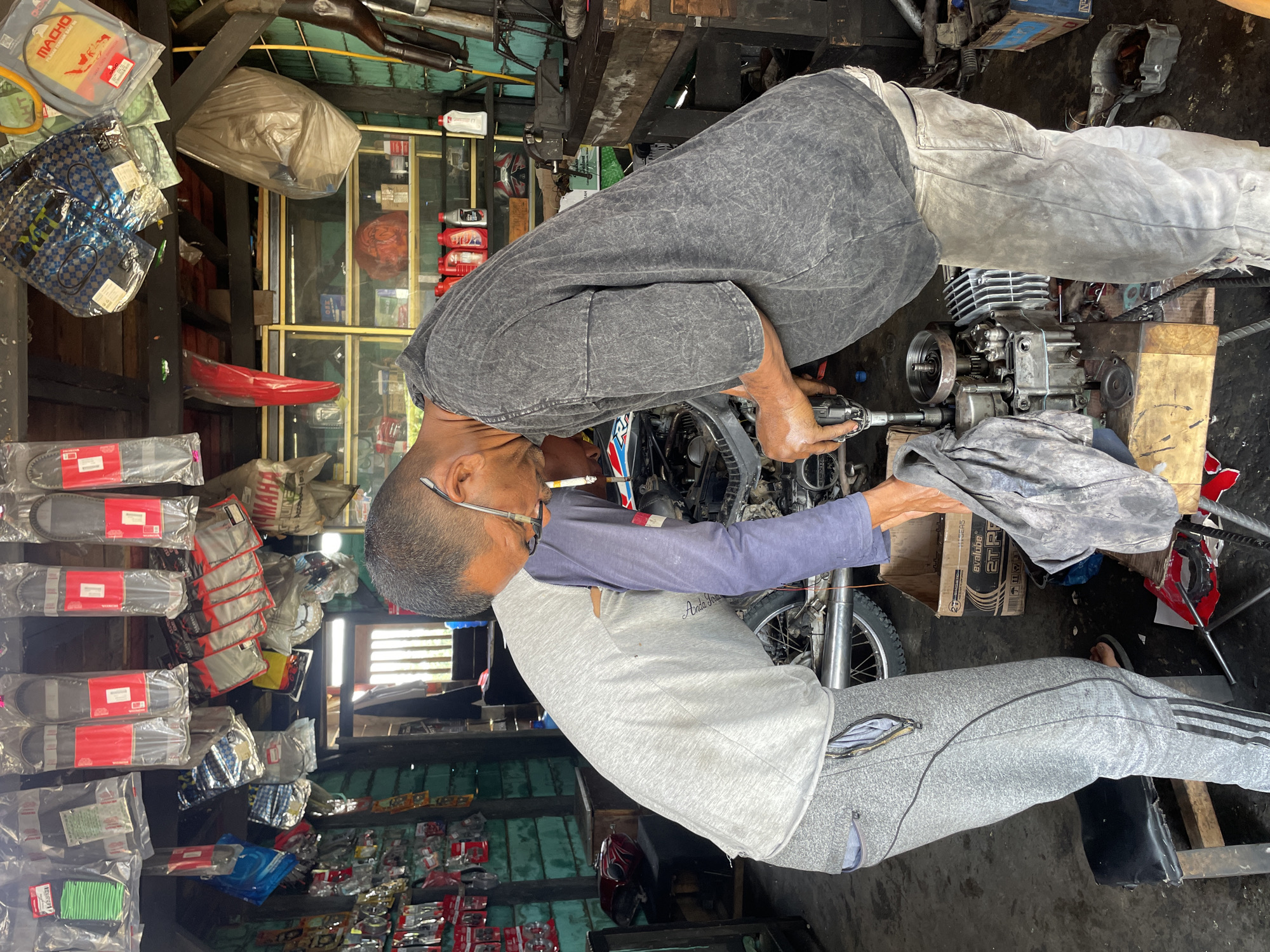 The image captures a bustling motorcycle repair shop in Glee Gogo, Indonesia, with two men focused on their work. In the foreground, a man wearing a grey t-shirt with blue long sleeves, grey sweatpants, and glasses observes or assists, with a cigarette in his mouth. His shirt bears the text "Arada Jeans." Behind him, another man in a dark grey t-shirt and light grey pants is actively engaged in repairing a disassembled motorcycle engine, holding a tool. The engine is positioned on a wooden block on the dark, utilitarian floor.
The shop environment is typical of a local garage, filled with various motorcycle parts and accessories. These items are stored on wooden shelves and hung around the space. Visible products include packaged drive belts, air filters, and bottles of engine oil. Text such as "MACHO," "YAMAHA," and "HONDA" can be seen on different packages. A cardboard box labeled "Eralube 2TR" is also present. The walls are a mix of wood and green corrugated material. The scene is illuminated by ambient daylight, indicating it is daytime. The overall atmosphere is one of practical, hands-on repair.