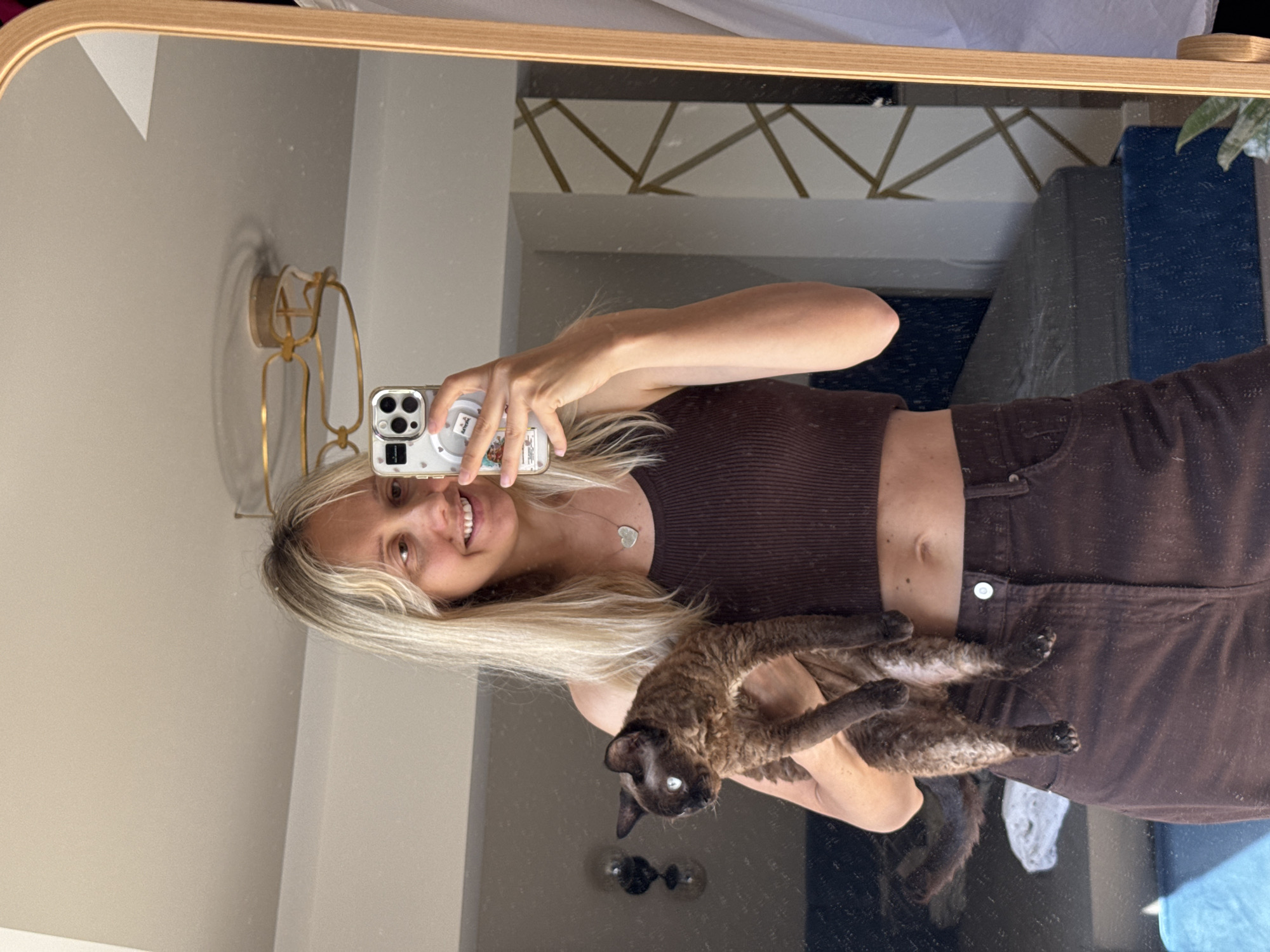 In a sunlit room, a blonde woman stands before a tall mirror, holding a dark, curly-haired cat. She wears a brown crop top and brown corduroy pants. The woman smiles at her reflection, holding up a smartphone to take a selfie. The mirror reflects the room behind her, showing a bed, a floor lamp, and a wall with a geometric pattern. The overall impression is one of casual domesticity. There is no indication of Carnikava, Latvia in the image.