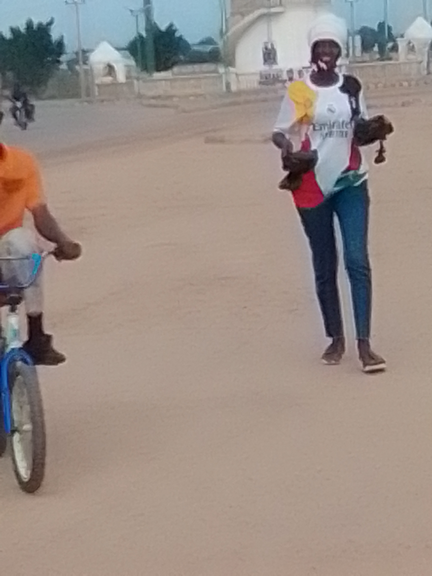 The image depicts a street scene in Kwadon, Nigeria. In the foreground, a person is riding a bicycle with a blue frame. They are wearing a bright orange t-shirt and dark pants. Further in the background, another individual is walking and holding several items, possibly for sale. This person is wearing a white t-shirt with a "Emirates" logo and colorful accents, a white head covering, and dark blue jeans. They are carrying two dark, textured objects, one in each hand, and what appears to be a yellow and red piece of fabric. The ground is sandy or dusty, indicative of a road in an outdoor setting. Behind the people, there are some buildings and trees, suggesting an urban or semi-urban environment. The lighting suggests it might be early morning or late afternoon.