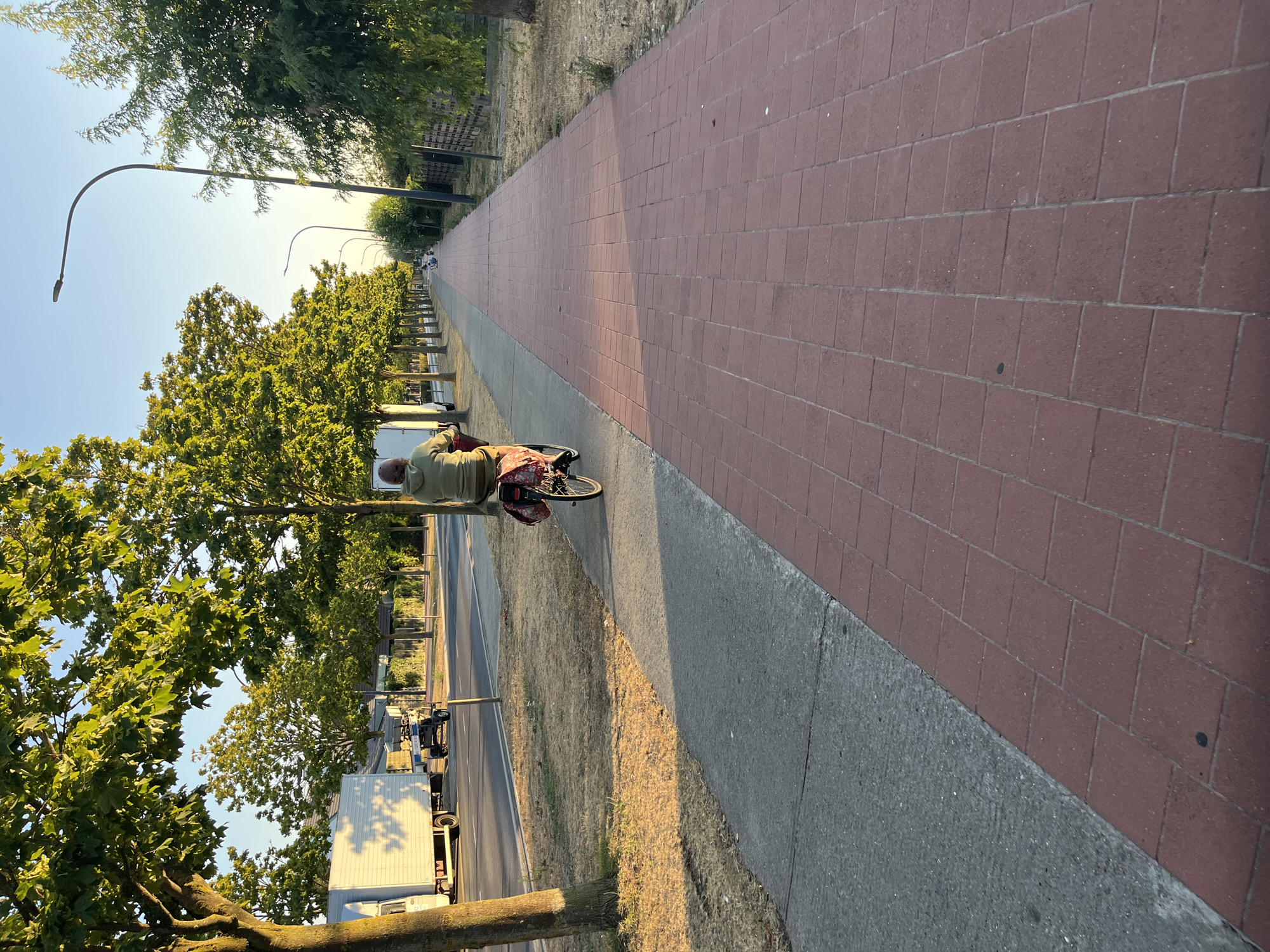 A person on a bicycle rides along a red brick pedestrian path next to a gray sidewalk. The path is lined with trees on the left and a fence and more trees on the right. The sky is clear and blue. The scene appears to be in a suburban area. The individual on the bicycle is wearing a light green hooded sweatshirt and is carrying a red bag on the back of their bike. There is a white truck and a small trailer on the road to the left of the sidewalk. The time of day appears to be late afternoon or early evening, given the long shadows.