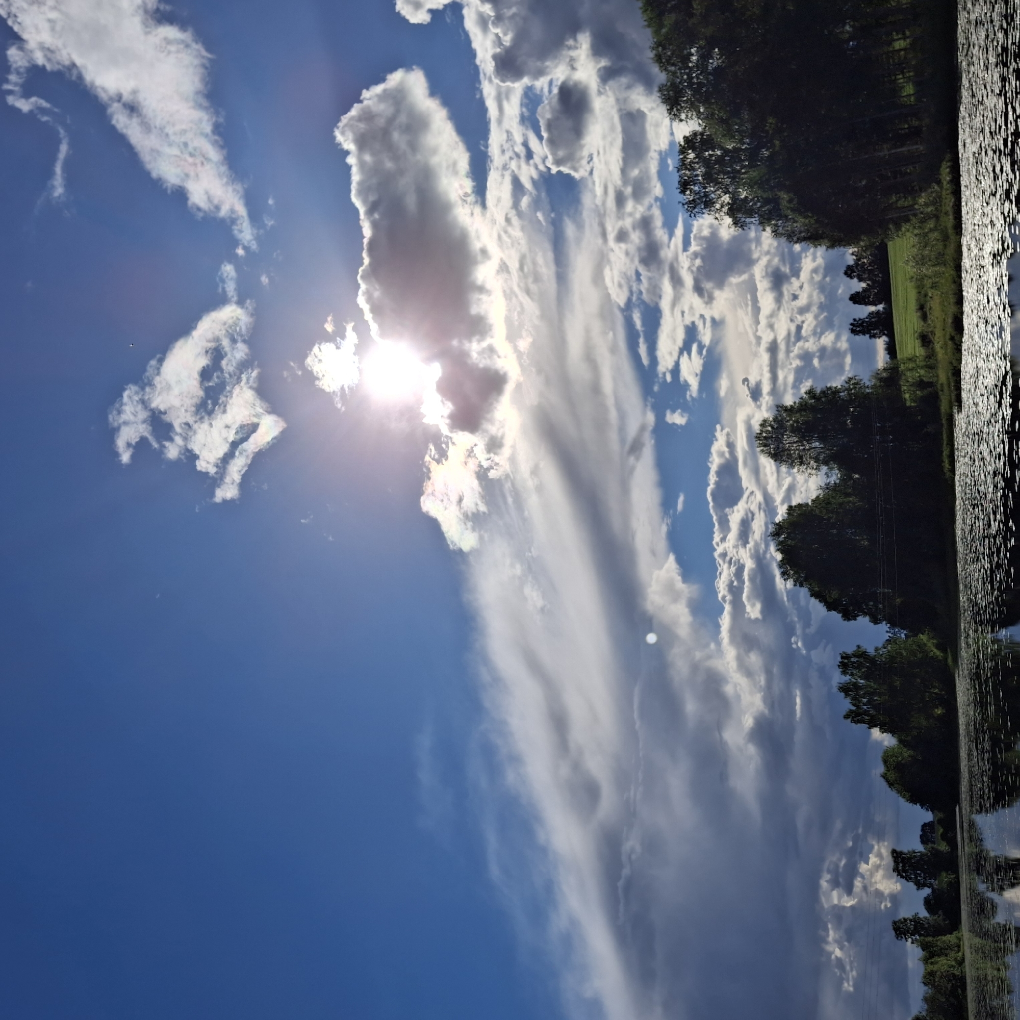 The image depicts a serene landscape scene. The dominant subjects are the sky, clouds, trees, and a body of water. The sky is a vibrant blue, adorned with a variety of clouds, ranging from wispy, streaky formations to larger, more voluminous cumulus clouds. The sun is visible, partially obscured by clouds, casting a bright glow and creating lens flare effects.
The scene transitions to a line of lush green trees that form a natural border along the edge of the water. The trees are densely packed and appear to be in full foliage, suggesting a warm season. Beyond the trees, a field of green grass can be seen, hinting at a rural or natural setting.
The lower portion of the image features a body of water, likely a lake or river, with a surface that is textured with ripples and reflections. The reflections of the trees and sky are visible on the water's surface, adding depth and visual interest to the scene. The overall impression is one of tranquility and natural beauty.
There is no visible text in the image. The time of day appears to be daytime, with the sun high in the sky. The weather seems pleasant, with a mix of sunshine and clouds. The location is suggestive of a natural environment, possibly a park or a nature reserve. The overall mood conveyed by the image is peaceful and contemplative.