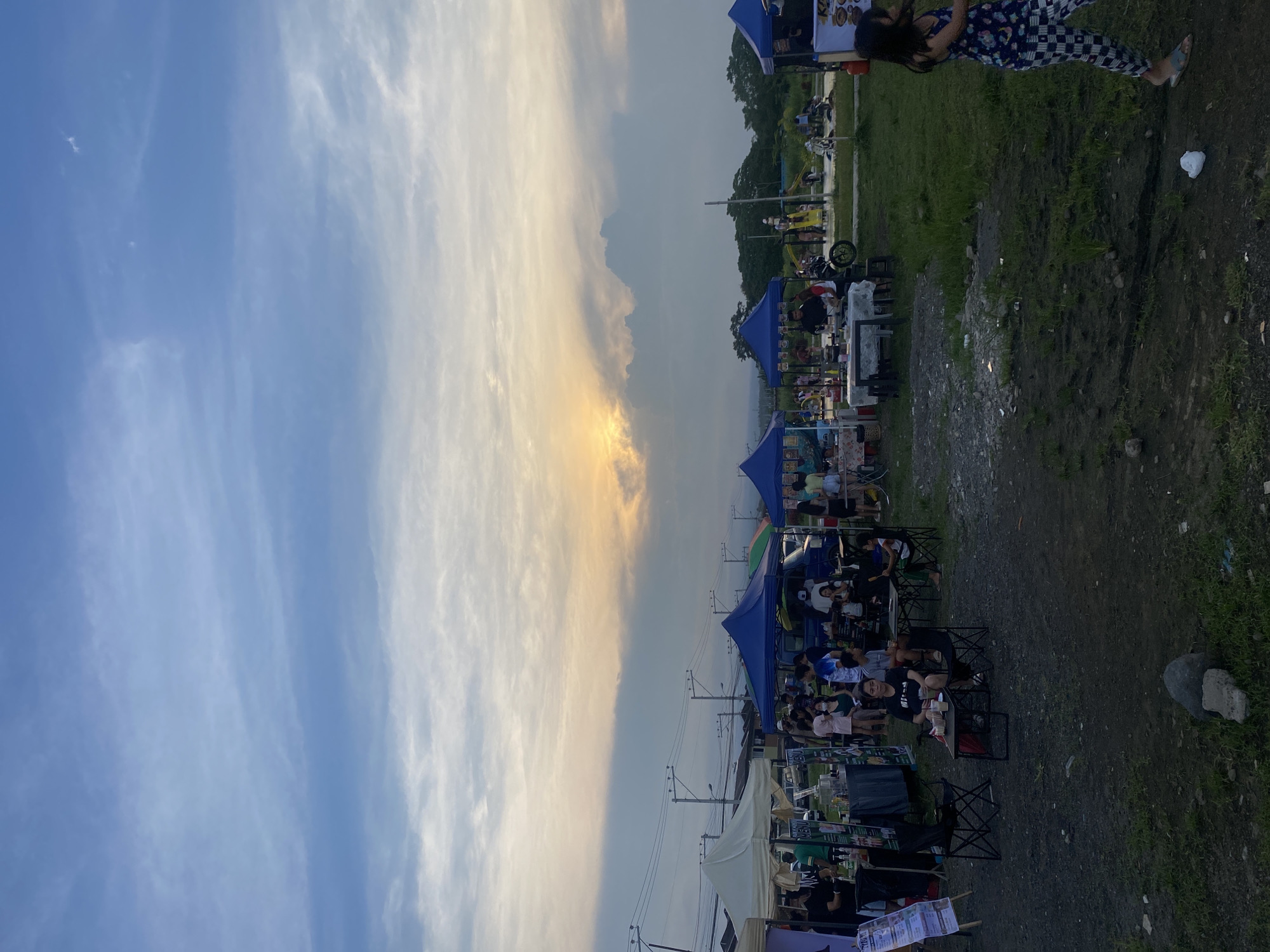 This image depicts an outdoor market or food park at sunset, likely in Davao City, Philippines. The scene is set along a wide dirt and gravel path lined with numerous blue and white canopy tents, functioning as market or food stalls.
The sky occupies the upper half of the image, showcasing a transition from a clear, light blue on the left to a warmer, orange-yellow hue on the right, illuminated by the setting sun behind scattered clouds.
People are the primary subjects, gathered in groups around foldable tables and chairs placed in front of the stalls. They appear relaxed, socializing, and enjoying food and drinks. A woman in distinctive checkered pants is partially visible on the far right. In the background, beyond the stalls and people, are trees and utility poles with overhead power lines. The foreground consists of the unpaved path, interspersed with patches of grass and small rocks, with a piece of white litter visible on the ground.
The activity is a casual gathering, suggesting an evening market or a food festival. The time of day is clearly late afternoon or early evening, characterized by the warm, dimming light of the setting sun. Visible text includes "COAL" on a banner towards the bottom left, and partial letters "Ki" and "O" on a banner in the upper right. The overall ambiance is social and leisurely.