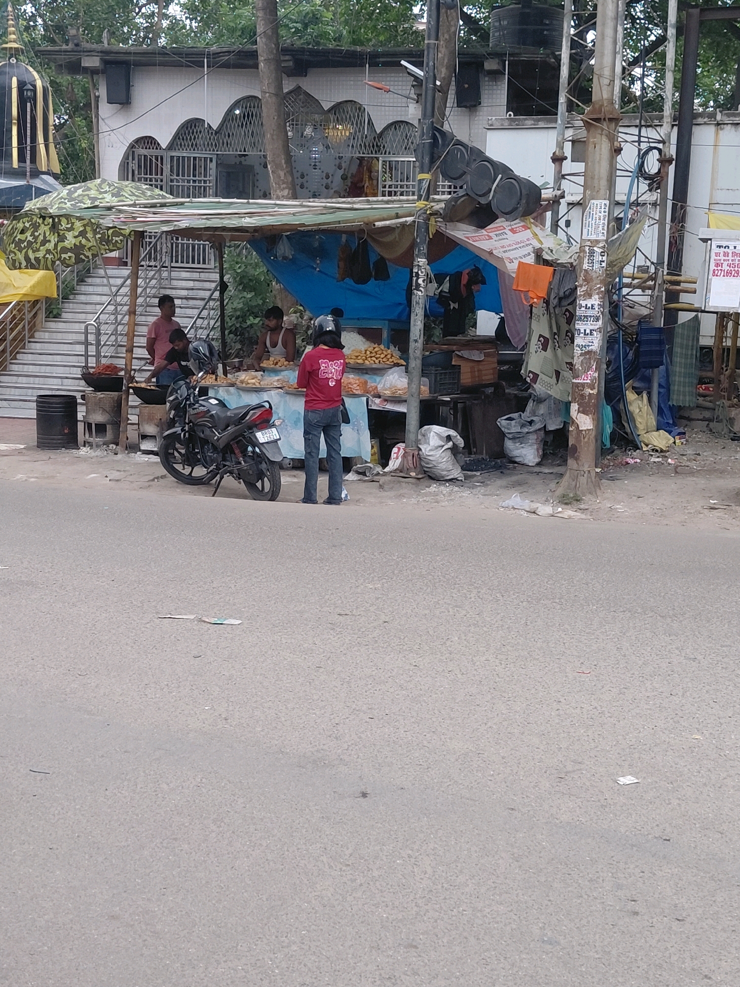 In Patna, India, a street scene unfolds with a food stall set up in front of a building with ornate arched windows and a prominent temple-like structure. A person in a red shirt and jeans stands near the stall, which is covered by a camouflage-patterned awning and a blue tarp. The stall displays a variety of food items, including what appears to be fried snacks and fruits. A motorcycle is parked to the left of the stall. In the background, a person wearing a tank top is visible behind the counter, seemingly serving customers or preparing food. The ground in front of the stall is littered with various items, including sacks and debris, suggesting a busy urban environment. Traffic lights hang from a pole, and various posters and advertisements are attached to it. The overall scene depicts a typical roadside food vendor in a bustling Indian city.