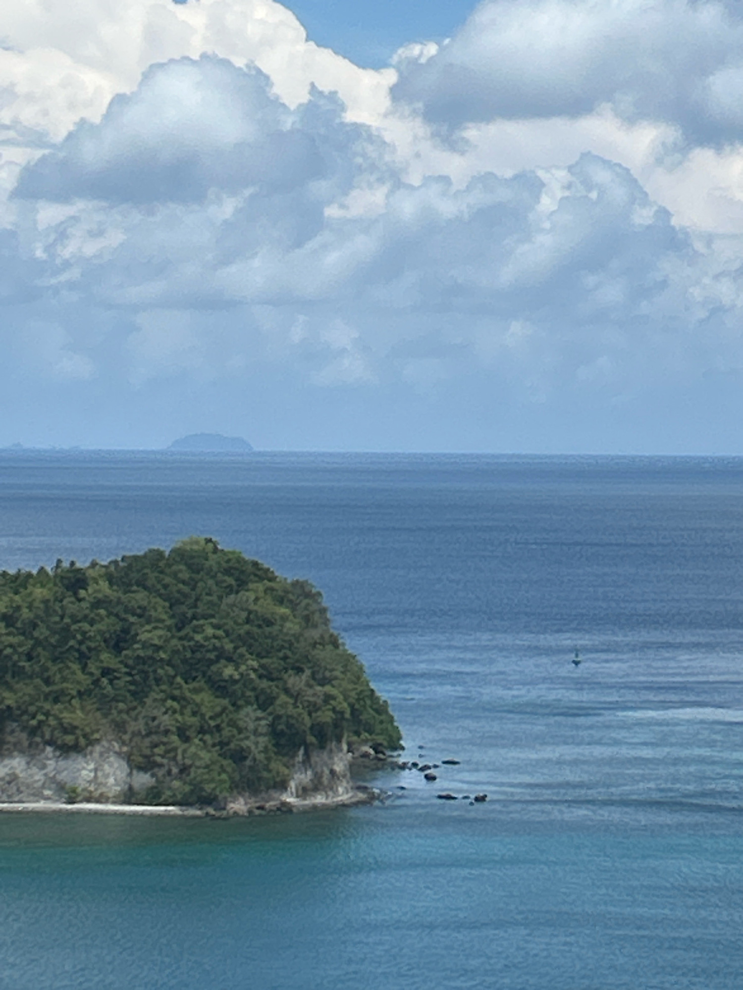 The image captures a serene seascape with a verdant island in the foreground, situated off the coast of Sabang, Indonesia. The island, covered in lush green foliage, slopes gently towards the water, revealing rocky outcrops and a small stretch of white sand. The crystal-clear turquoise waters surrounding the island give way to deeper blue expanses further out. In the distance, a hazy outline of another island is visible on the horizon, under a vast sky filled with dramatic, billowing clouds against a backdrop of pale blue. A solitary sailboat can be spotted in the middle ground, adding a touch of human presence to the natural landscape. The overall scene is one of tranquility and natural beauty.