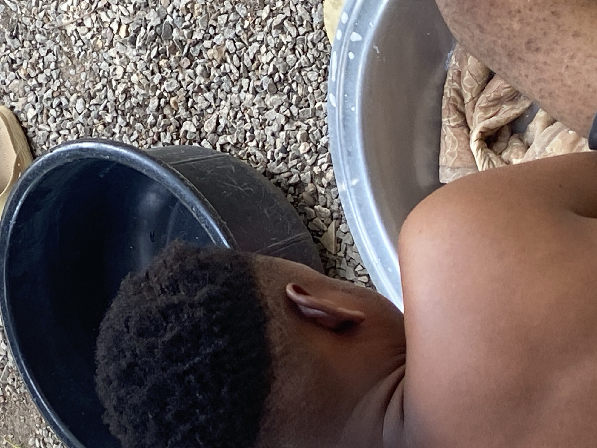 A child is being bathed outdoors. The child is sitting in a black plastic tub filled with water. To the right of the child, there is a light gray plastic tub that contains a patterned cloth or blanket and water. The background is gravel ground cover. The photo was taken in Mararaba, Nigeria.