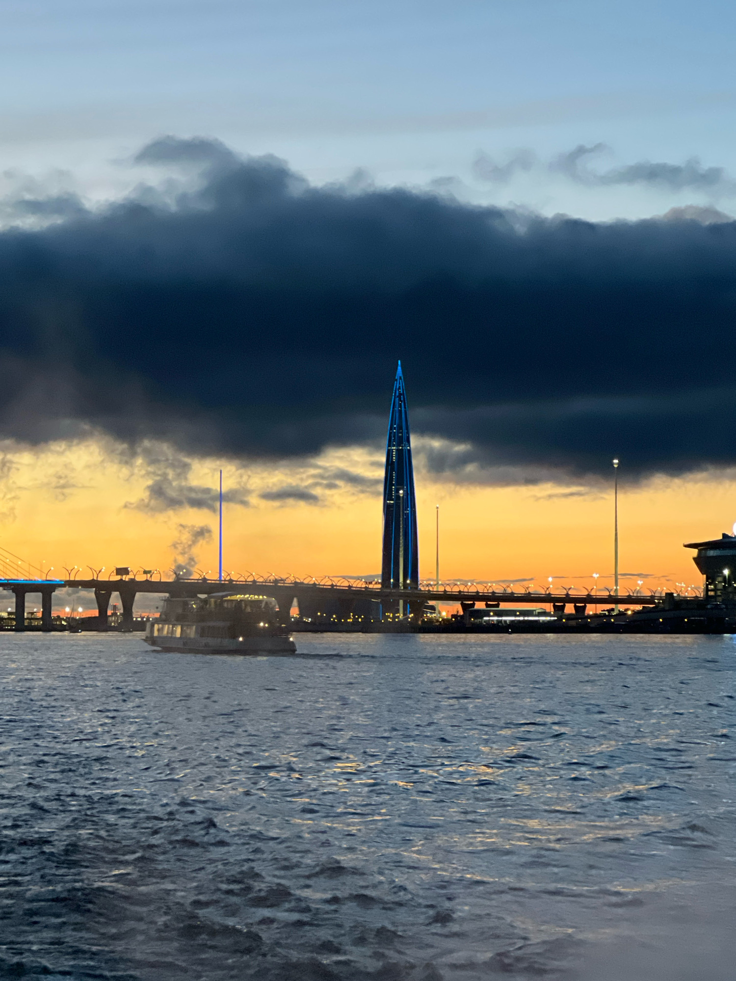 The image depicts a view of the Gulf of Finland in Saint Petersburg, Russia, at dusk. The foreground shows choppy water with reflections of light. In the midground, a tour boat is navigating across the water. Behind the boat, a modern bridge, likely part of the Western High-Speed Diameter, spans the water. A distinctive illuminated skyscraper, the Lakhta Center, is visible in the background, its blue lights contrasting with the orange and dark blue hues of the sunset sky. Dark, dramatic clouds hang over the scene, adding to the atmospheric quality of the image.