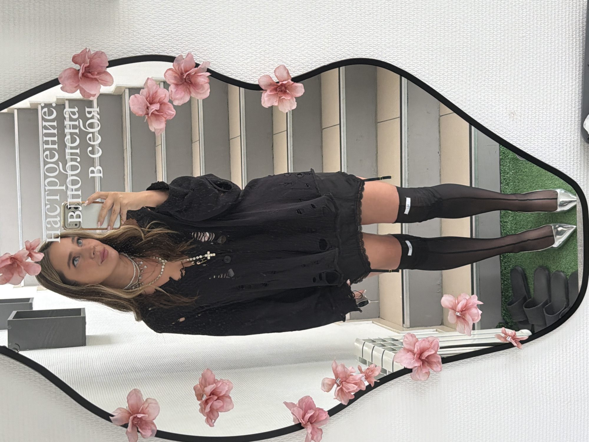 A young woman is featured in a mirror selfie, holding a smartphone to capture her reflection. The mirror has an irregular, undulating black frame and is decorated with numerous pink artificial flowers along its edges. The woman is dressed in a distressed black top or short dress, complemented by black sheer thigh-high stockings with a notable white band, and pointed silver heels. Her outfit is accessorized with several layered necklaces, one featuring a prominent cross pendant. She has long, wavy light brown hair and looks slightly upwards and to the side, away from the phone's lens, with a contemplative expression.
The reflection in the mirror reveals a background of grey and white stairs, suggesting a multi-level indoor or semi-outdoor setting within a modern building. A patch of green, resembling artificial grass, is also visible at the bottom right of the reflection, where a pair of black sandals or slippers rests. The mirror itself is mounted on a white, textured wall. Along the upper left side of the mirror, white Russian text is inscribed vertically, reading "настроение: влюблена в себя," which translates to "mood: in love with myself." The scene appears to be well-lit, indicating daytime. This setting is located in Chaikovsky, Russia.