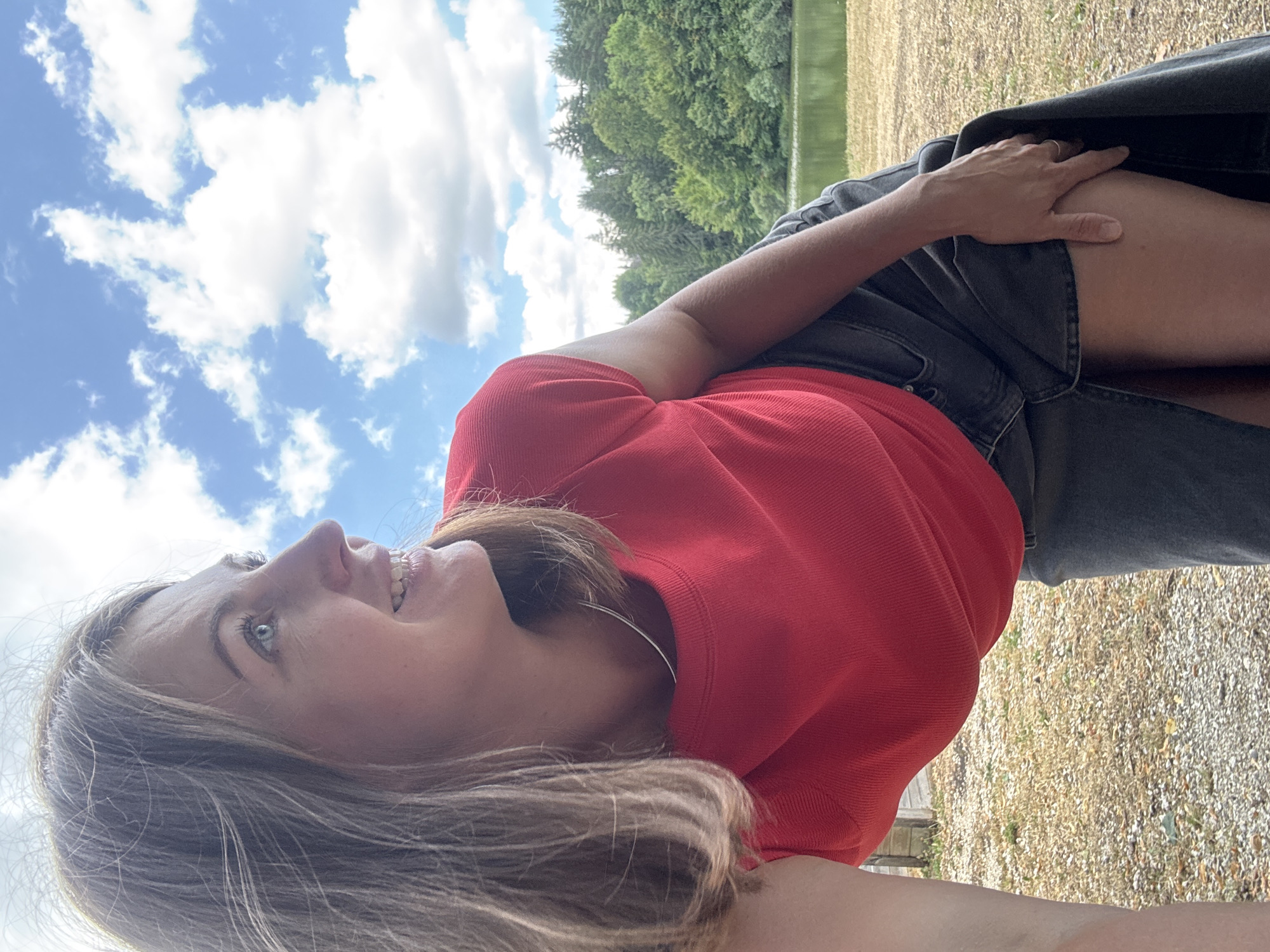 A woman with blonde hair is captured in a medium-shot from the chest up, looking to her right. She is wearing a red t-shirt and a dark denim skirt. Her head is tilted back, and she is smiling, revealing dental braces. The background features a blue sky with scattered white clouds, a body of water, and a dense forest. The foreground shows dry, gravelly ground. The location appears to be a park or natural area, potentially near Coteaux du Lizon, France, given the lush greenery and the presence of a lake or river.
