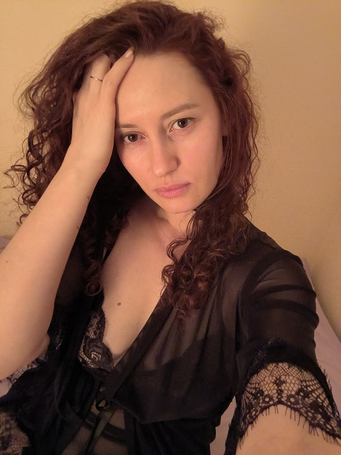 An adult female is depicted in a close-up indoor portrait, facing directly towards the camera. She has long, curly, reddish-brown hair and a light complexion. Her left hand is raised, with fingers resting on her forehead. A thin, plain ring is visible on the ring finger of her left hand. She is wearing a sheer black robe or negligee with lace detailing on the sleeves, over a black lace undergarment. A small mole is visible on her upper chest. The background consists of a plain, light-colored wall, with a partial view of light purple fabric on the right side, suggesting a private room setting. The lighting is warm and soft. No specific geographic location within Sankt-Peterburg, Russia, can be determined from the visual information provided.