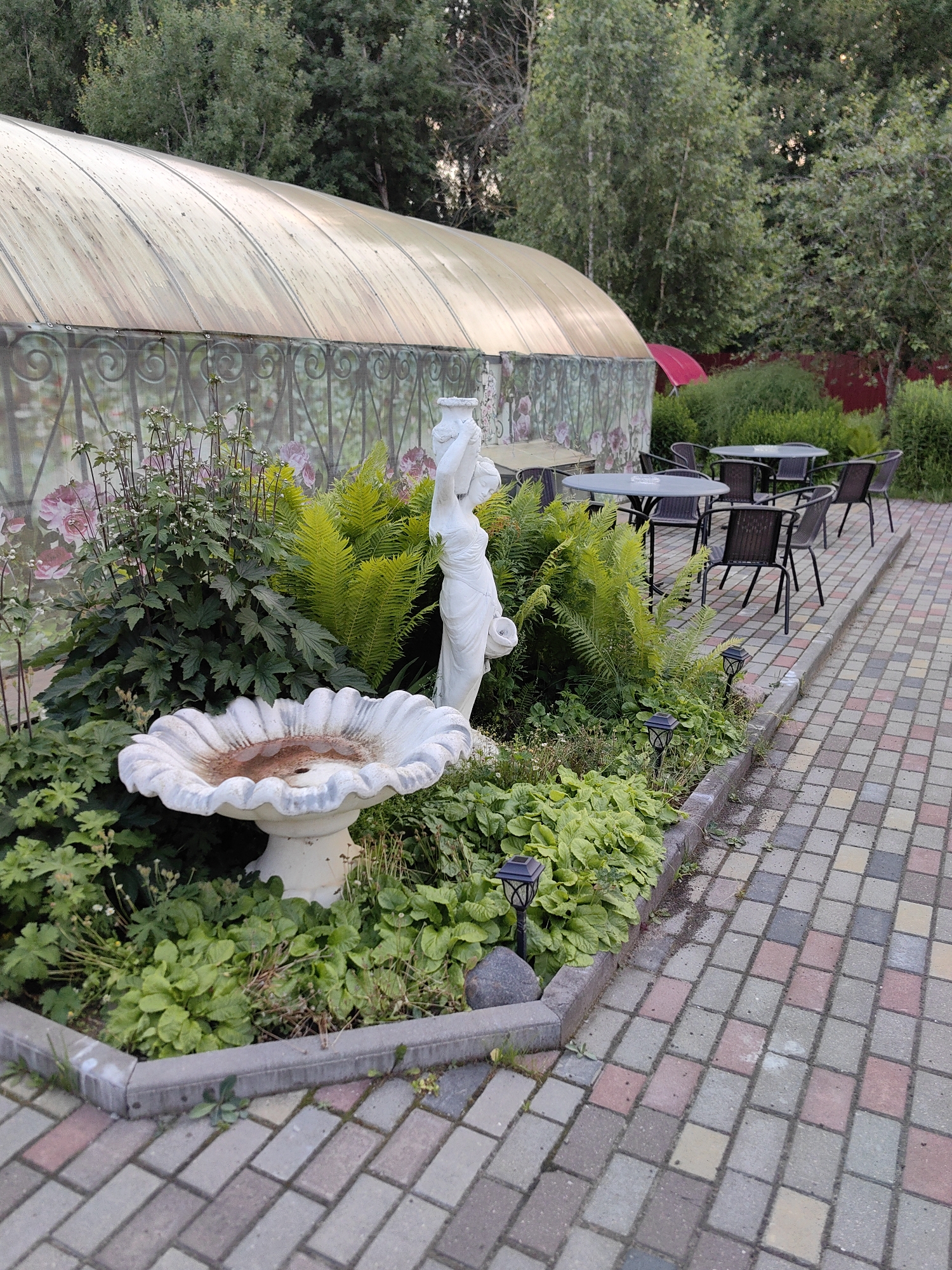 In the city of Фаніпальскі сельскі Савет, Belarus, an outdoor seating area is depicted. A patterned brick walkway lines the foreground, leading towards several round tables with dark chairs. Behind the tables, a garden bed features a white, ornate birdbath as its centerpiece, surrounded by lush green foliage, ferns, and a white statue of a woman. A greenhouse with a translucent, curved roof stands in the background, partially obscured by trees and a red umbrella. The scene is set during daylight hours, with natural light illuminating the area.