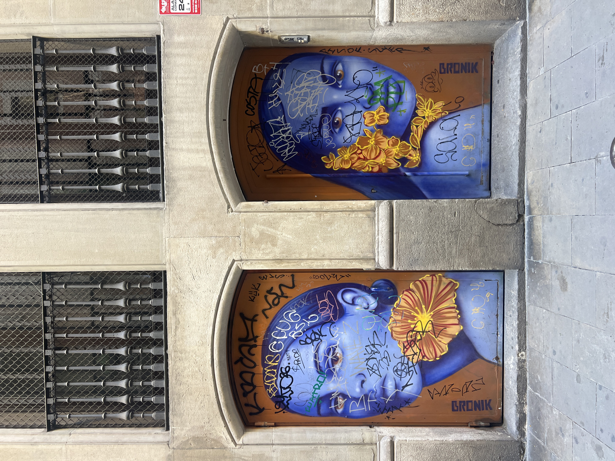 The image captures a segment of an urban building facade in Barcelona, Spain, centered on two adjacent arched doorways and two rectangular windows positioned directly above them. The building's exterior is constructed from light beige, textured stone or stucco.
Each arched opening contains a brown metal or wooden door, both heavily adorned with large-scale street art depicting female faces rendered in shades of blue. The artwork is extensively overlaid with numerous graffiti tags and scribbles in black, white, and green. On the left door, a blue-skinned woman with dark hair and light brown eyes gazes slightly right, with a large orange and yellow flower painted near her chin. The right door features a similar blue-skinned woman, facing slightly left, with a cluster of yellow flowers and green foliage painted over her left cheek. The signature "GRONIK" is vertically visible on the lower outer edges of both doors.
Above the doorways, two recessed windows are visible. Each window is protected by a dark grey wrought iron railing, composed of eight decorative vertical balusters, and a fine mesh screen positioned behind the railing. To the right of the right window, a red and white rectangular sign displaying "ALARMA 24H," "INSP. SERVICIOS CONECTADOS 24H," and a local Barcelona phone number (932 44...) is affixed to the wall. A small grey rectangular intercom or doorbell unit is mounted on the wall to the right of the right doorway. The foreground consists of a sidewalk paved with grey square stone tiles. The scene is illuminated by natural daylight.