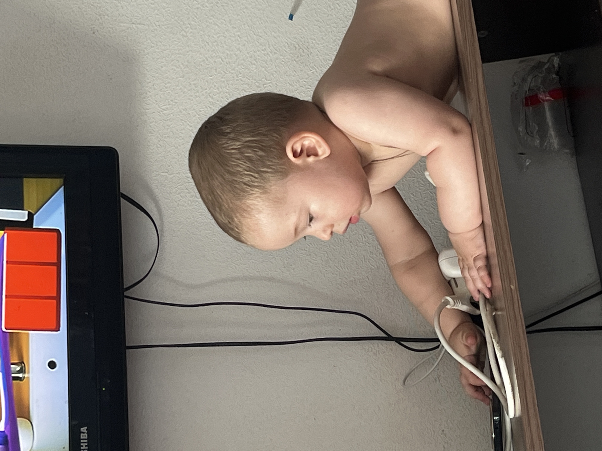 The image shows a young child, likely a toddler, leaning over a shelf. The child has light brown hair and is looking down at something on the shelf. They are shirtless. On the shelf, there are several electrical cords and a white power adapter. A television is visible in the background, displaying a colorful, animated image. The wall behind the television appears to be textured. The overall scene suggests a domestic environment, possibly a living room. No specific details in the image allow for the determination of its location within Chelyabinsk, Russia.