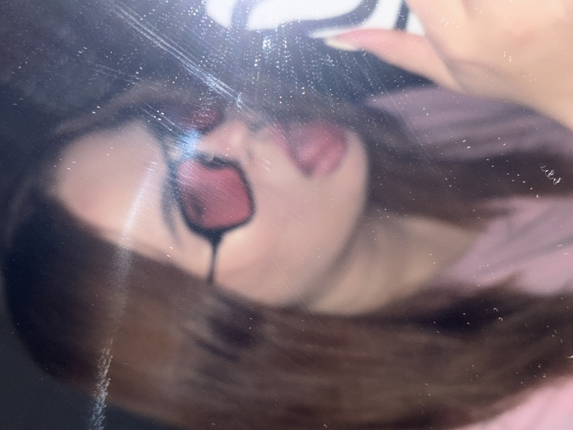 The image shows a person with red, cartoonish makeup resembling a heart on their face, partially obscured by a blurry reflection, possibly on a glass surface. The person has dark hair and is wearing a pink garment. A hand holding an object, possibly a phone case with a cartoon design, is visible at the top. There is no indication of the location being Krasnodar, Russia. The focus is on the person and the reflection.