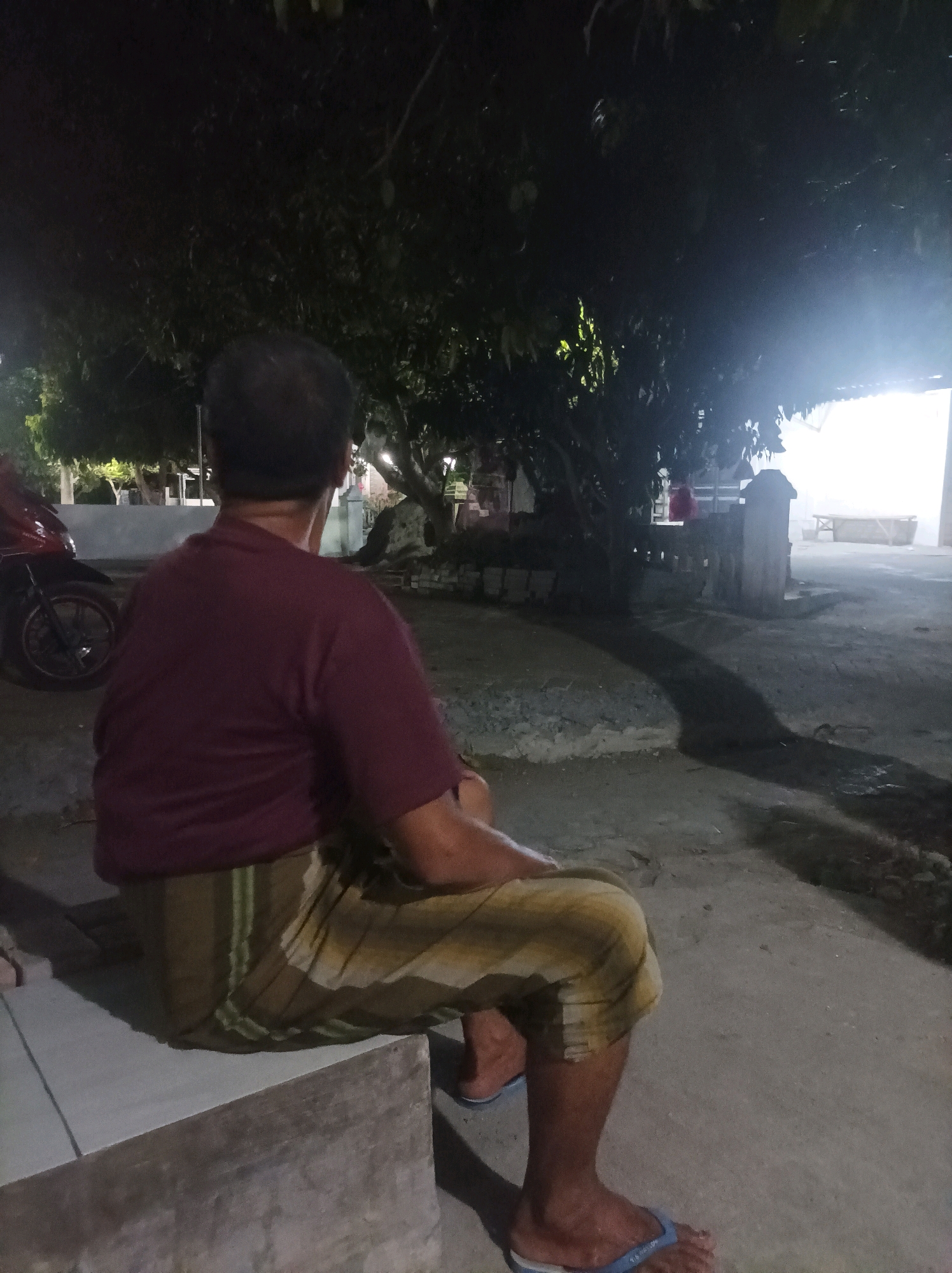 In the city of Canga'an, Indonesia, a man is seen from behind, sitting on a low concrete structure. He is wearing a dark maroon t-shirt and a sarong with a patterned design in yellow, brown, and green stripes. His feet are clad in blue flip-flops. The setting appears to be outdoors at night, with a large tree casting a shadow across the ground. In the background, a motorcycle is parked to the left, and a building with bright lights can be seen to the right. The ground is paved with concrete, and there are some low walls and other structures visible in the distance.