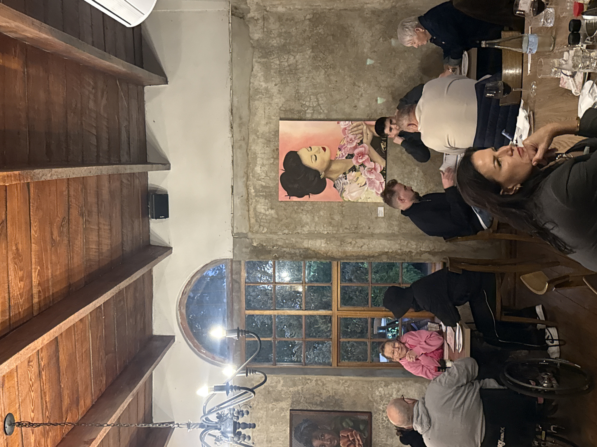 The image shows a group of people gathered around a table, seemingly enjoying a meal. The setting appears to be an indoor dining area, possibly in the city of Paarl, South Africa. The room has exposed wooden beams on the ceiling, a large window with a curved top, and concrete or stone walls adorned with paintings. The table is set with plates, glasses, and bottles, indicating a formal or semi-formal meal setting. The people present are of various ages, engaged in conversation. One individual appears to be in a wheelchair.