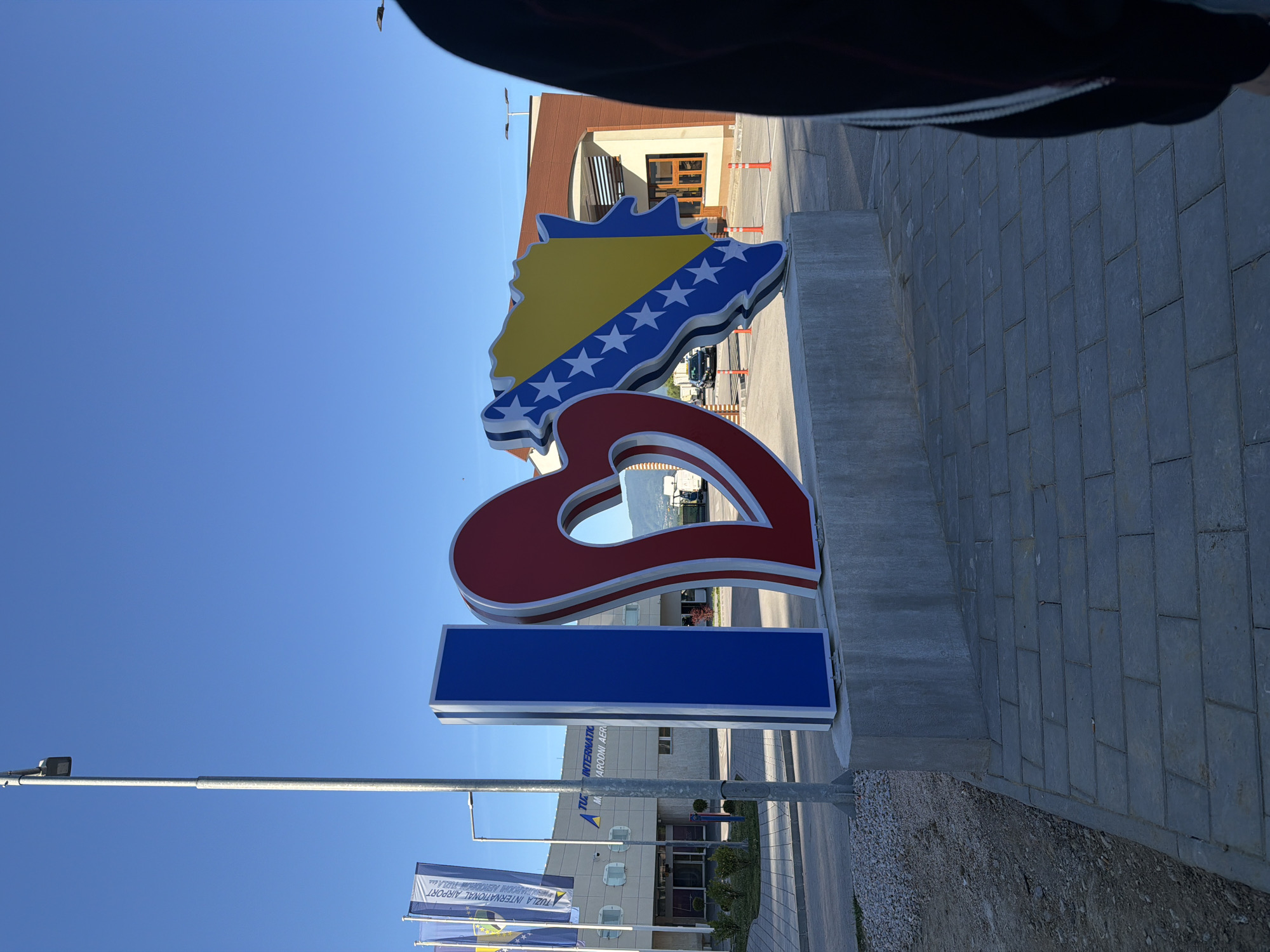 The image depicts a sign in Gornje Dubrave, Bosnia and Herzegovina, likely near the Tuzla International Airport, as suggested by the airport flags and building in the background. The sign consists of the shape of Bosnia and Herzegovina colored in the national flag colors, a red heart, and a blue rectangle. The sky is clear and blue. A person is partially visible in the upper portion of the frame. The ground is paved with gray stones.