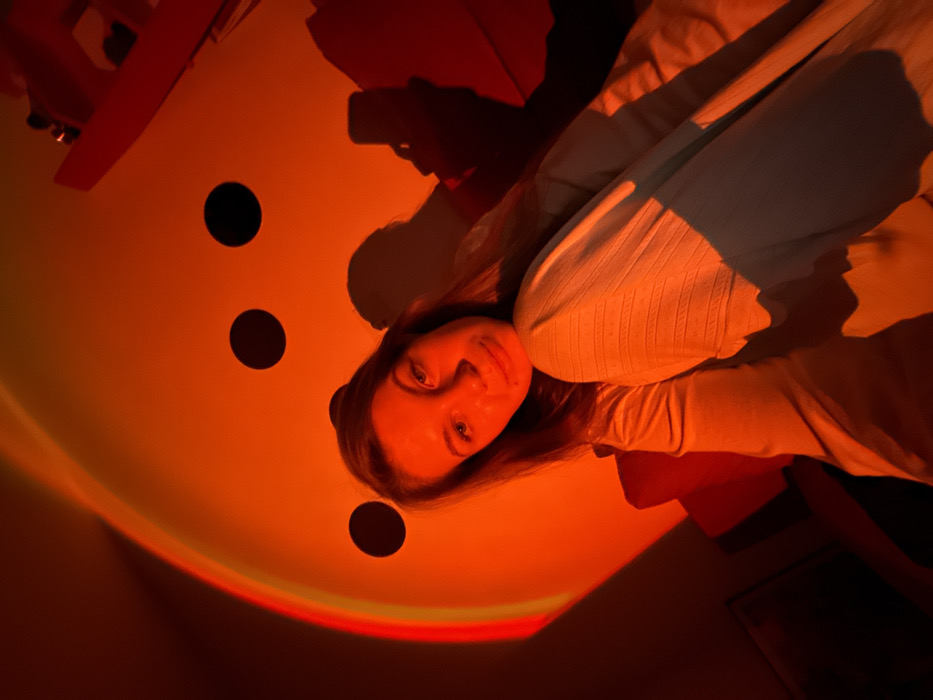 The image depicts an indoor scene located in the city of Vozdovac Urban Municipality, Serbia. A female individual is positioned in the foreground, seated, with her upper body and face illuminated predominantly by intense red and orange artificial light. She is looking directly towards the camera. She wears a light-colored long-sleeved top and ribbed light-colored pants; her left knee is bent and elevated, with her left arm resting on it. The background features a large, curved, light-colored wall surface, partially adorned with three dark circular spots, exhibiting a gradient of yellow, orange, and red artificial illumination on its left side. A dark shadow depicting a hand holding a smartphone is cast on the wall to the right of the individual, indicating the camera's proximate position. Partial views of dark furniture or objects are present in the lower-left and upper-right corners of the frame.