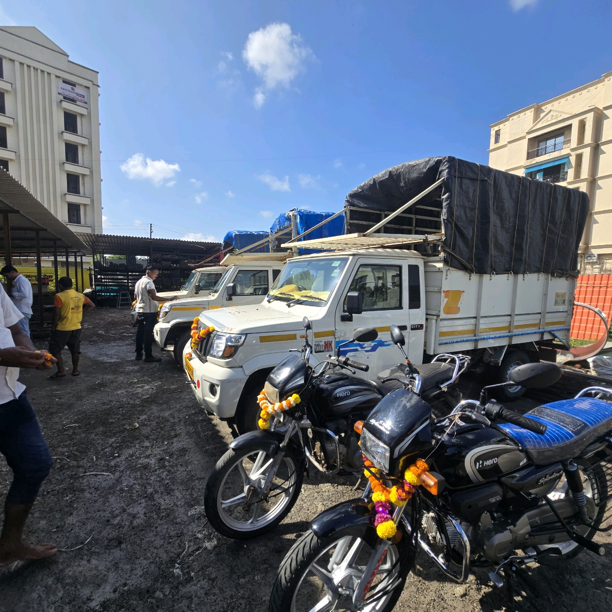 In Palghar, India, a row of vehicles is parked on a dirt lot. Three white trucks with covered beds are positioned side-by-side. Two black motorcycles are parked in front of the trucks, each adorned with garlands of yellow and pink flowers. The environment suggests an outdoor marketplace or storage area, with a building visible in the background. Several individuals are present, some standing near the vehicles and others appearing to be involved in activity around the trucks. The sky is blue with scattered clouds.