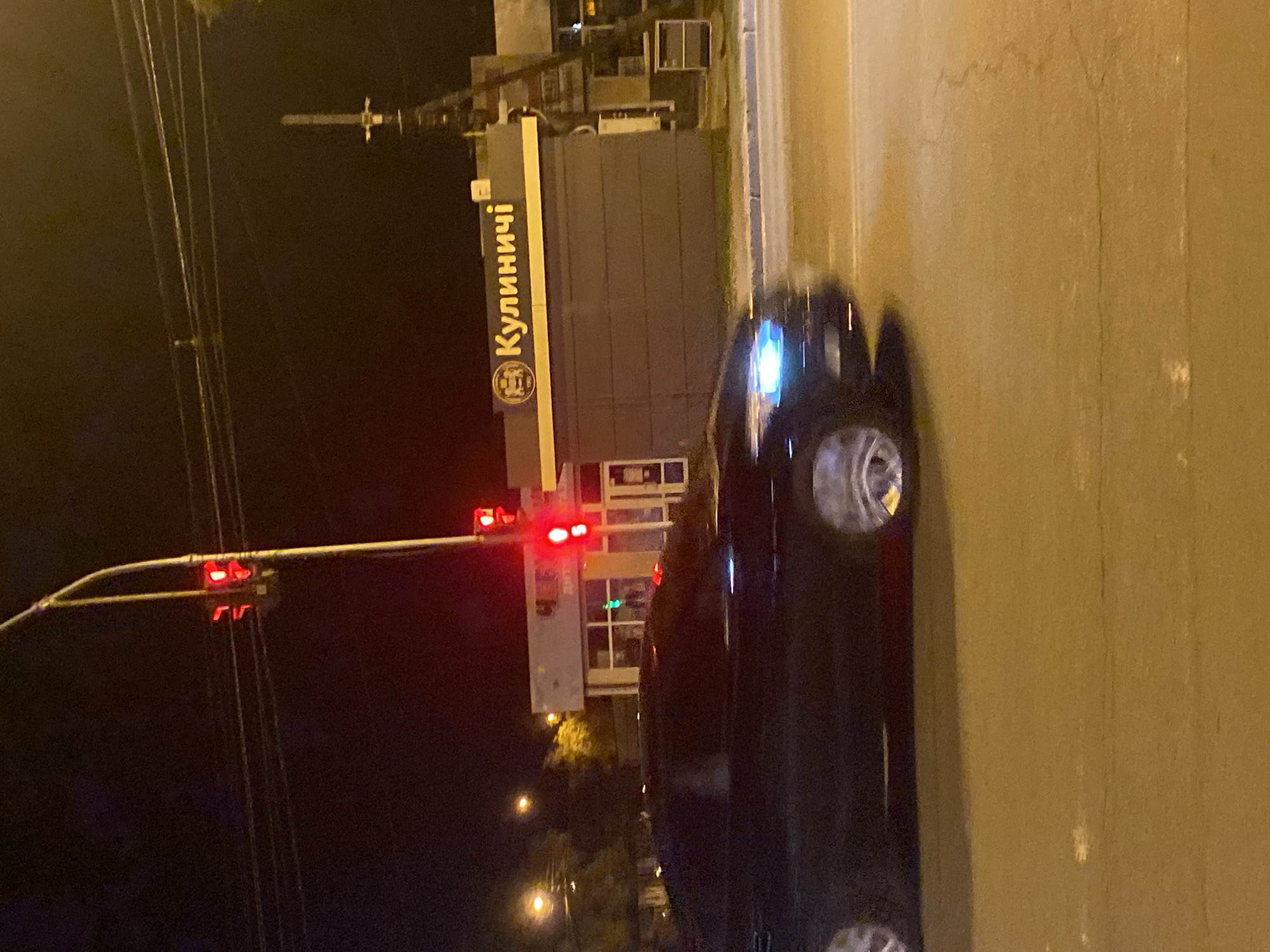 The image depicts a street scene at night in Poltava, Ukraine. A dark-colored car is in motion on the right side of the frame, its headlights blurred due to the movement. To the left, a traffic light shows a red signal. Behind the traffic light and the car is a building with the word "Кулиничі" displayed in Cyrillic script. Power lines are visible in the dark sky above the traffic light. The street appears to be illuminated by streetlights and the lights from the building.
