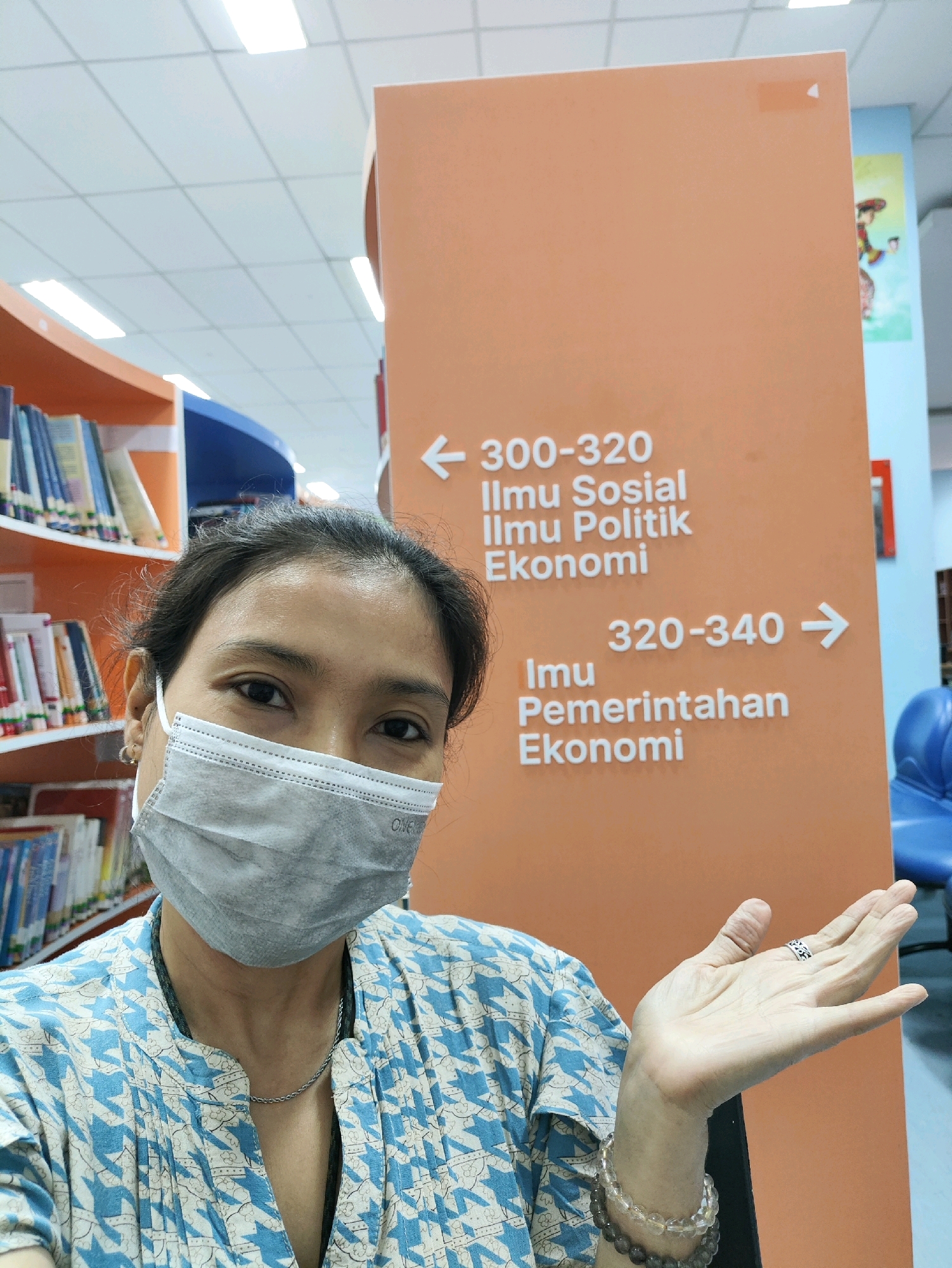 In a library setting in East Jakarta, Indonesia, a woman wearing a face mask and a blue and white patterned shirt is visible. She is standing in front of an orange sign with white text and arrows. The sign indicates sections for "Ilmu Sosial Ilmu Politik Ekonomi" (Social Science, Political Science, Economics) from 300-320 and "Ilmu Pemerintahan Ekonomi" (Government Science, Economics) from 320-340. Bookshelves filled with books are visible on the left side of the image. The woman is holding her left hand up, showing a ring and a bracelet. The environment appears to be well-lit, with fluorescent ceiling lights.