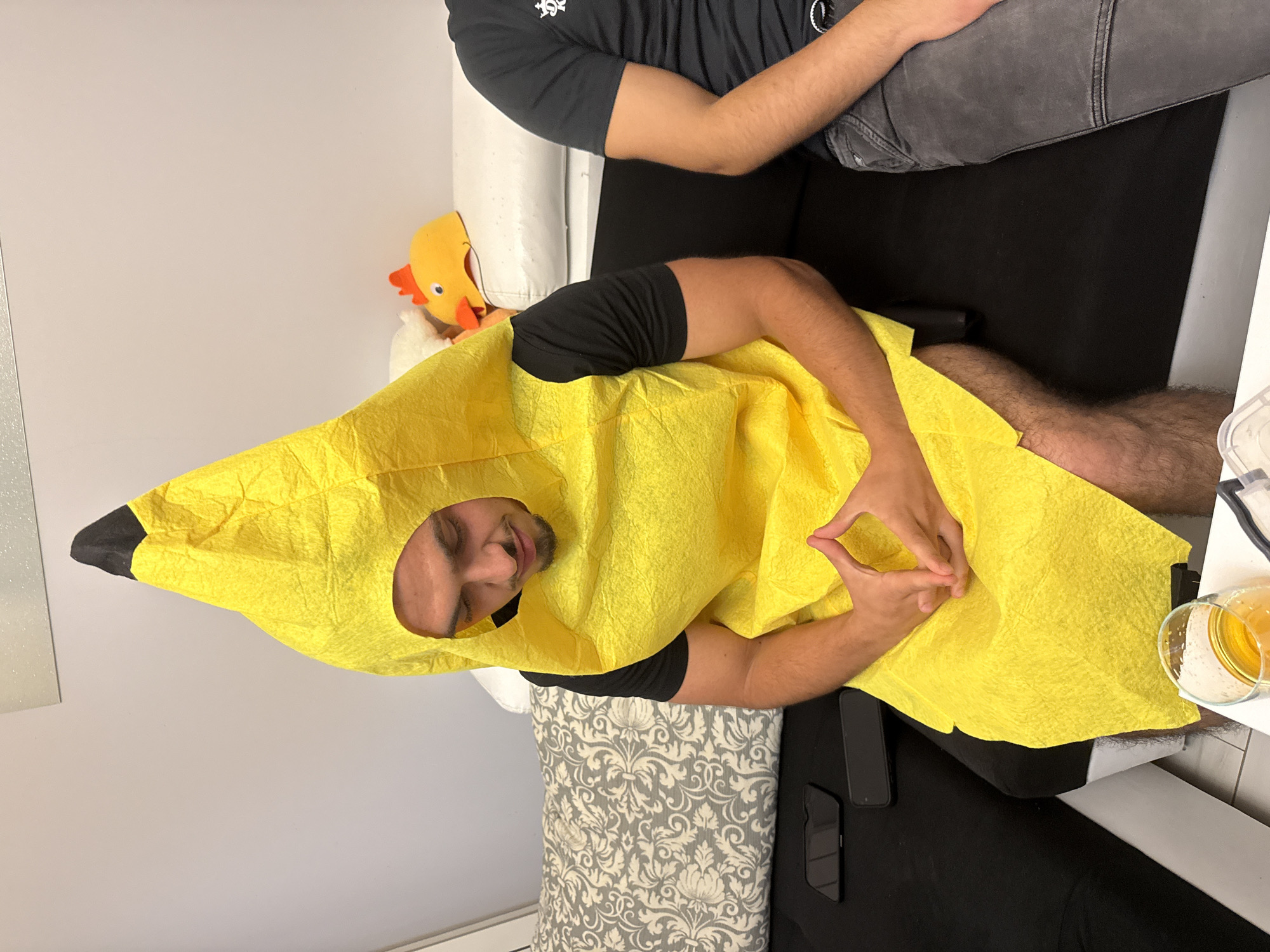 An image, rotated 90 degrees clockwise, captures a casual indoor scene in Sofia, Bulgaria, featuring two men. The primary subject is a man lying on his back, dressed in a bright yellow banana costume with a black tip. He wears a black t-shirt underneath the costume, and his bare legs are visible. He has a beard and a slight smile or smirk on his face, looking towards the camera. His left hand is raised, with fingers forming a distinct triangular or pyramid-like gesture.
Another man is partially visible in the upper part of the frame, sitting upright behind the costumed man. He is wearing a dark grey t-shirt and grey jeans. The setting appears to be a relaxed living room or bedroom. Behind the head of the man in the banana costume, a small yellow chicken plush toy with a red comb peeks out from behind a white sofa or bed.
On a dark surface to the right of the costumed man, two black smartphones are placed. Nearby, on a white surface, rests a clear glass containing an amber-colored liquid. The furniture includes a white sofa or bed, accompanied by a grey and white patterned cushion or pillow. The background consists of plain light-colored walls and a reflective surface, possibly a mirror, on the left. The lighting is artificial and consistent with an indoor setting, with no specific cues to the time of day. No visible text is present in the image.