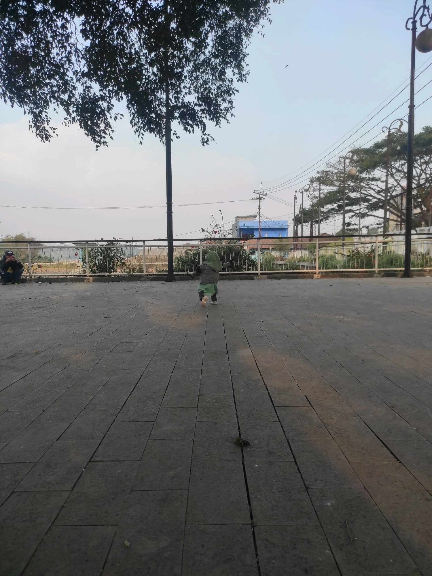 In Ciburuy, Indonesia, a child wearing a green hijab and a grey sweater walks away from the camera across a paved plaza. To the left, a person wearing a black baseball cap and a dark hooded jacket sits on the ground, looking at a phone. In the background, a white metal railing separates the plaza from a strip of green vegetation and some buildings. Overhead, a large tree branch with dark leaves extends into the frame from the upper left. Power lines and streetlights are visible in the distance. The sky is pale blue.