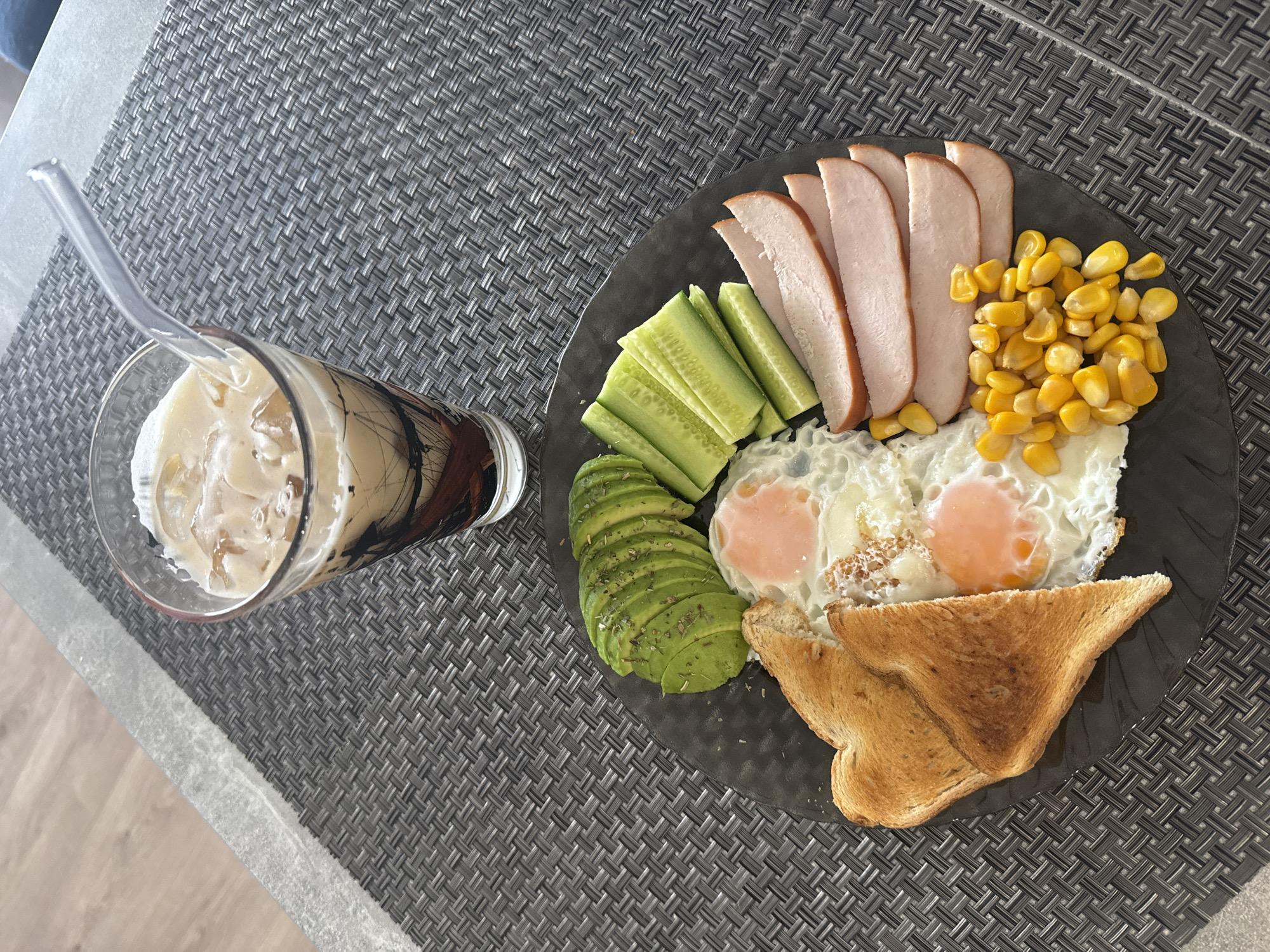 The image depicts a plate of food and a drink on a gray woven placemat. The plate contains sliced turkey breast, cucumber sticks, sliced avocado, fried eggs, toasted bread, and corn kernels. The drink is a glass with ice and a milky liquid, with a straw. The location is not discernible and cannot be determined to be Izhevsk, Russia, from the image alone.